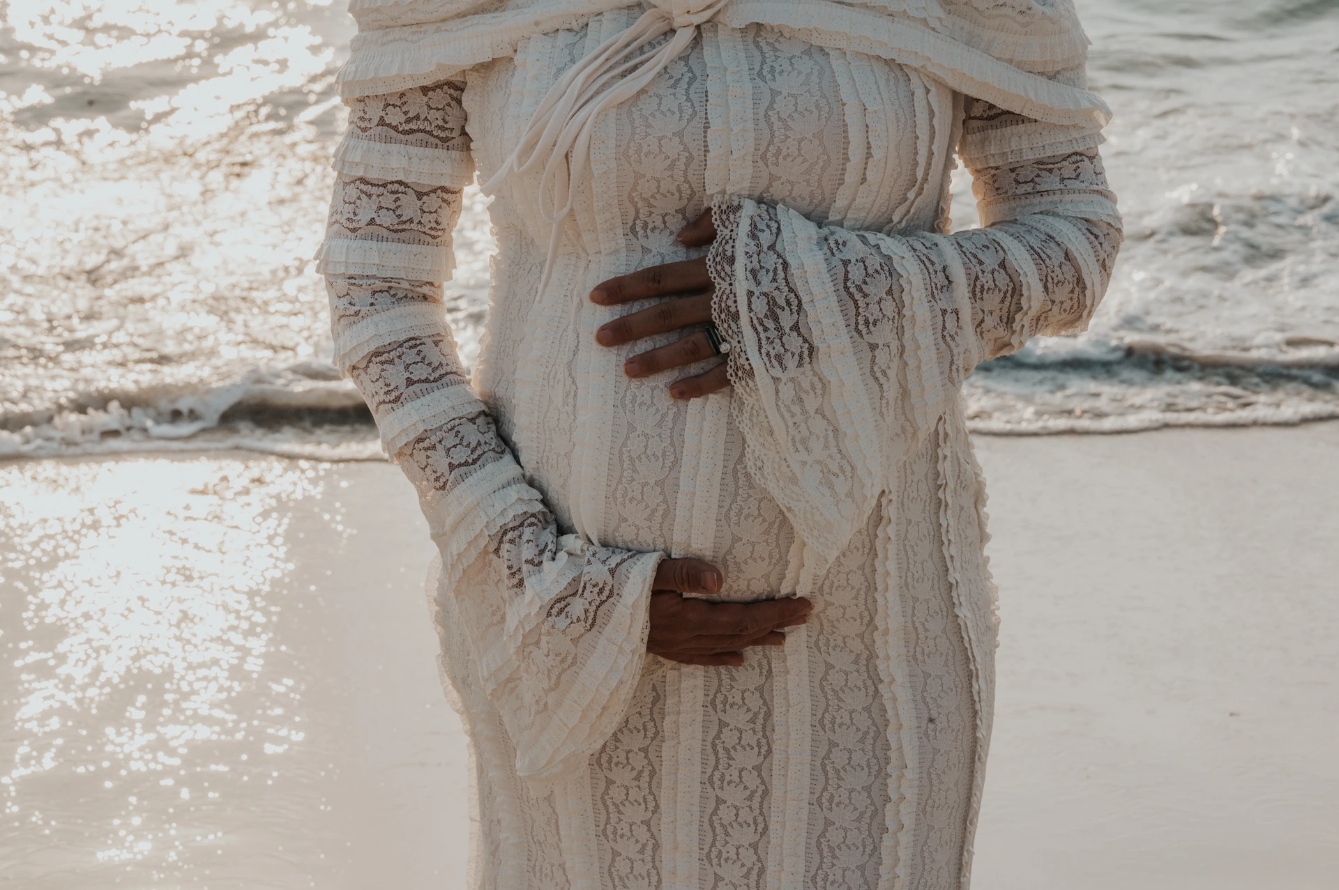 A pregnant woman in a lace dress holds her bump beside sunlit waves during a close-up Dubai beach maternity portrait.