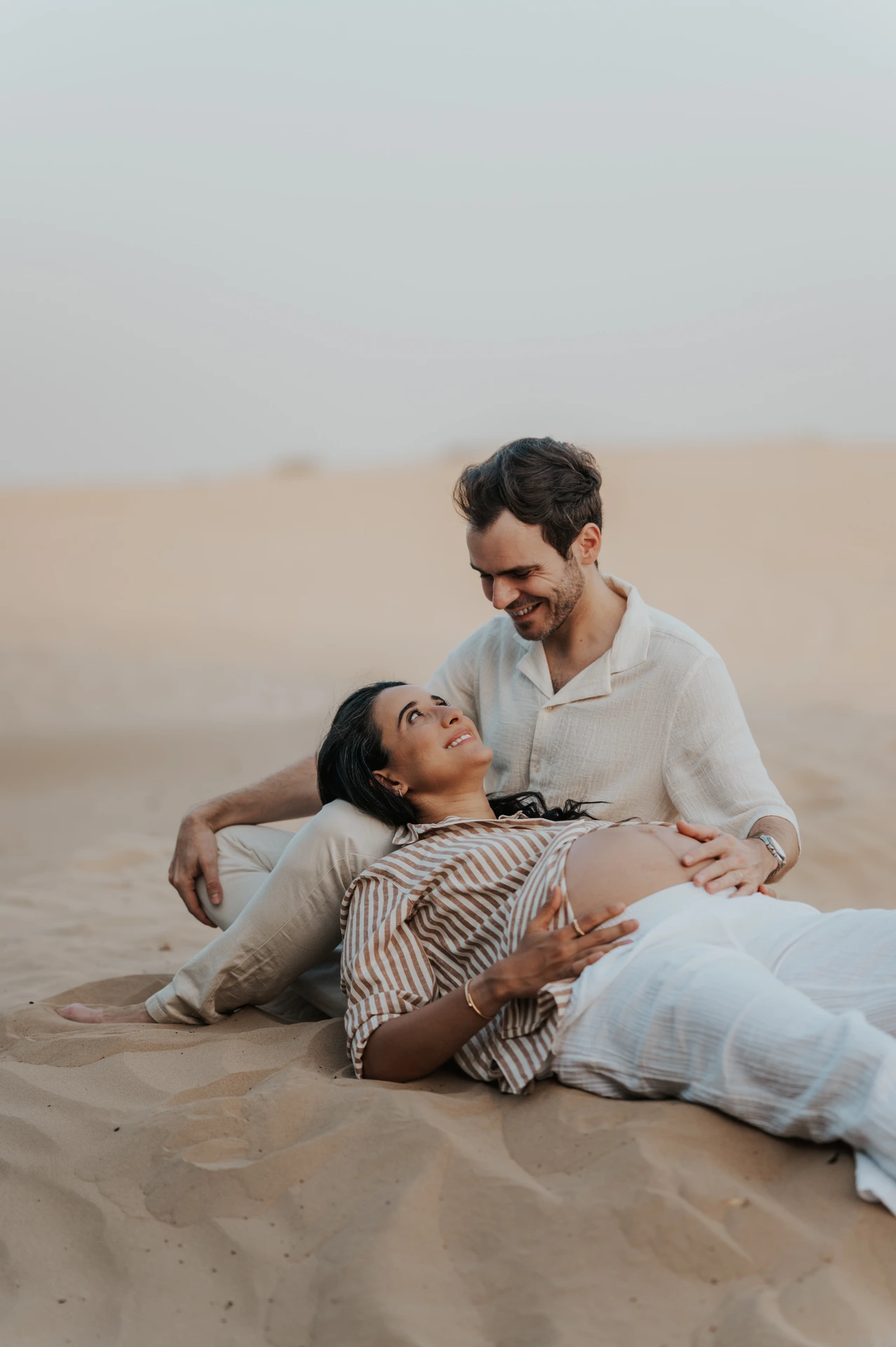 An expectant couple reclines on a Dubai desert dune, smiling at each other during their warm-toned maternity photoshoot.