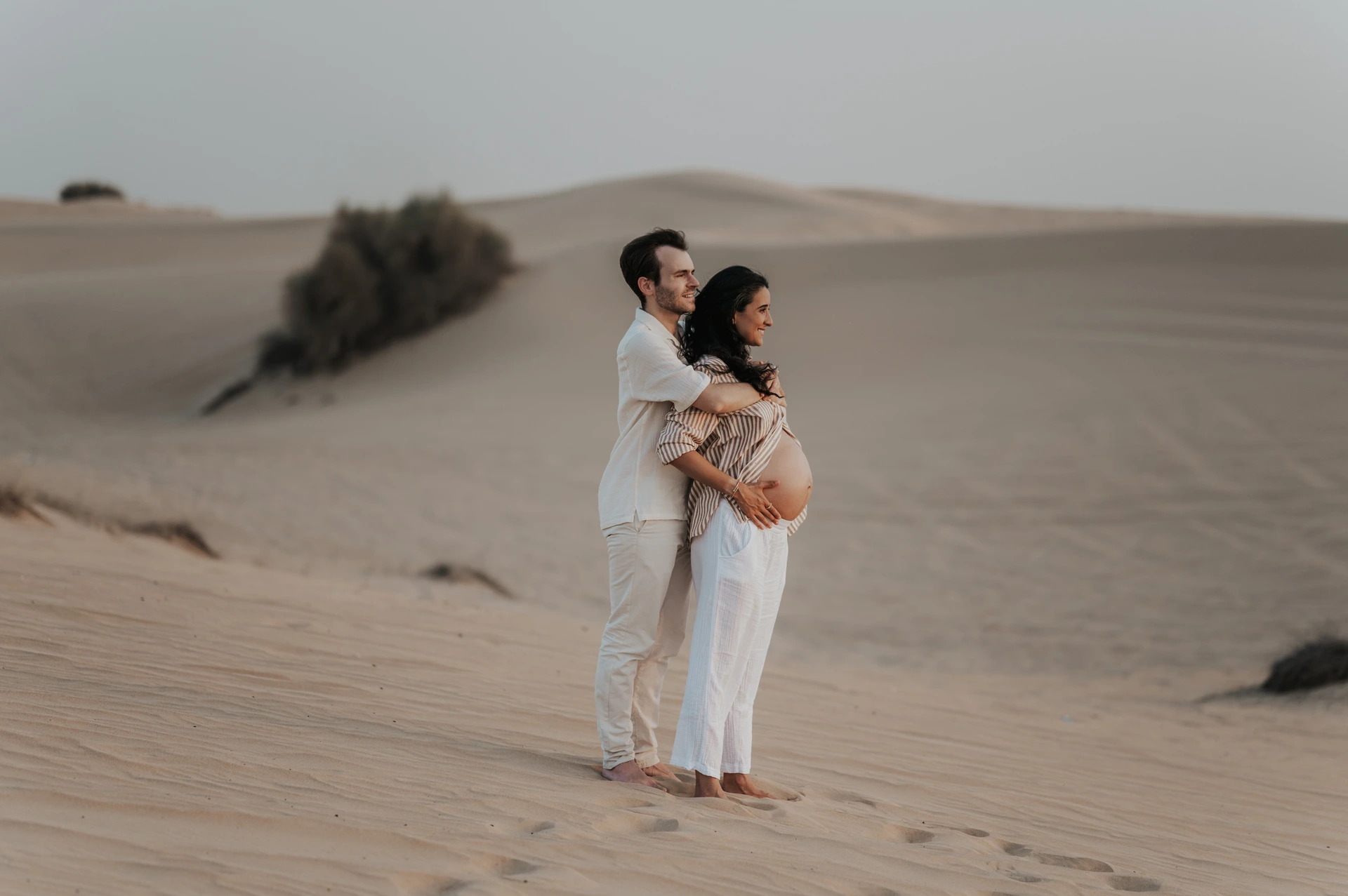 Dubai Desert Maternity Couple Standing on Dunes A pregnant woman stands in front of her partner as he embraces her on a wide Dubai desert dune during their maternity session.