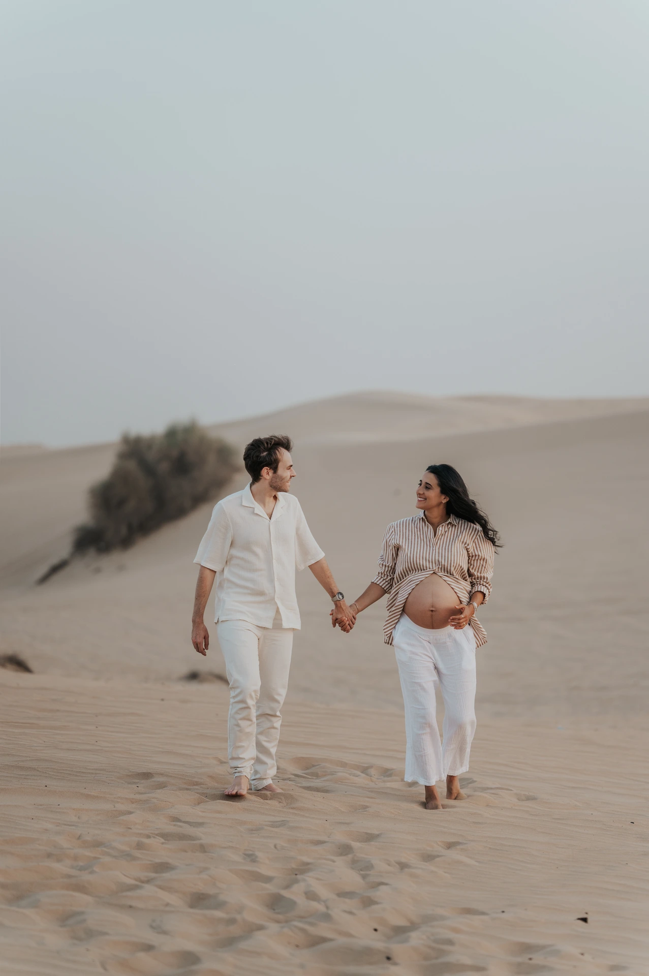 An expectant couple walks barefoot hand in hand across a Dubai desert dune during their sunset maternity photoshoot.