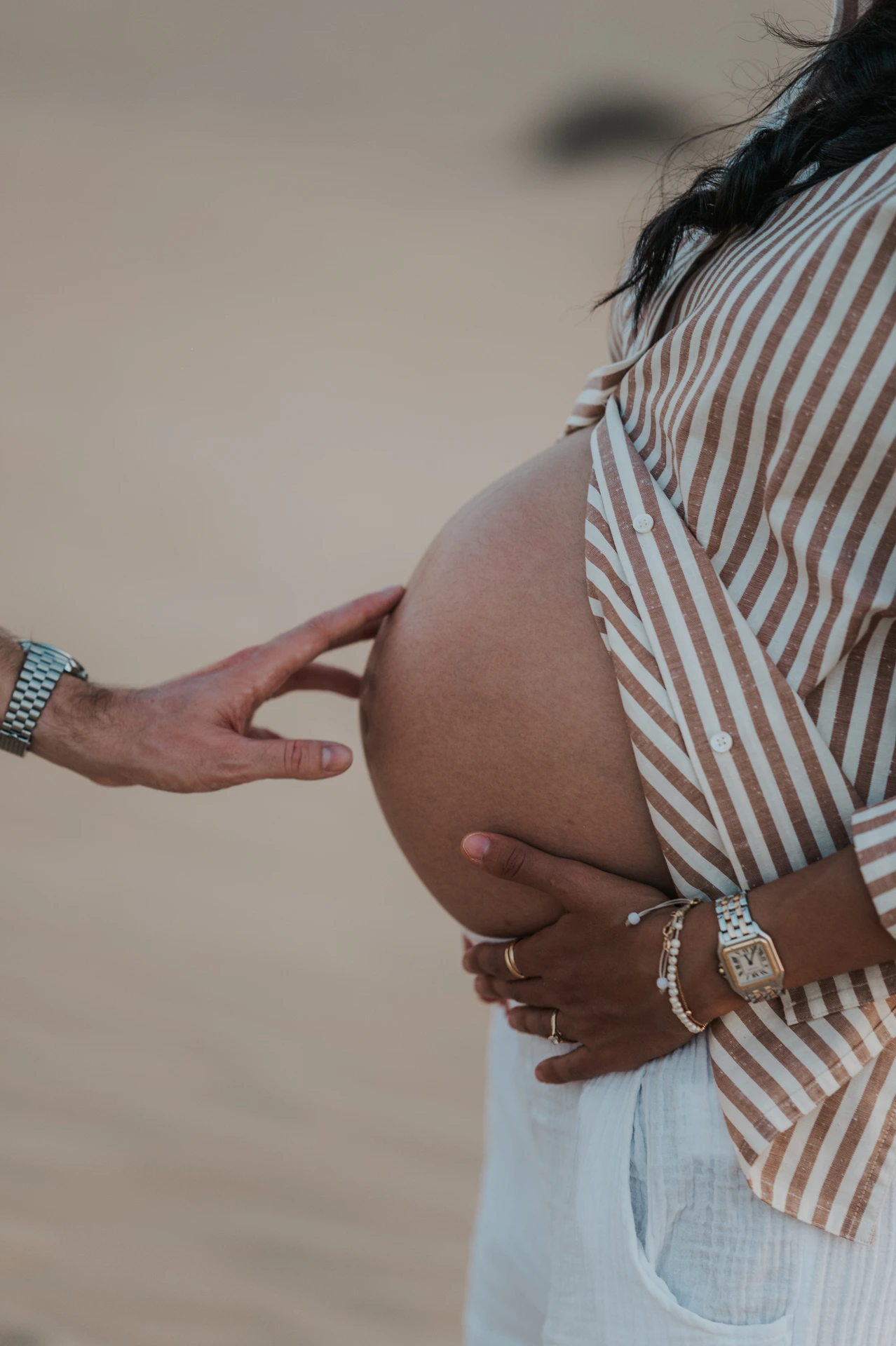 A partner reaches toward a pregnant woman's bump while she holds it on a Dubai desert maternity shoot.