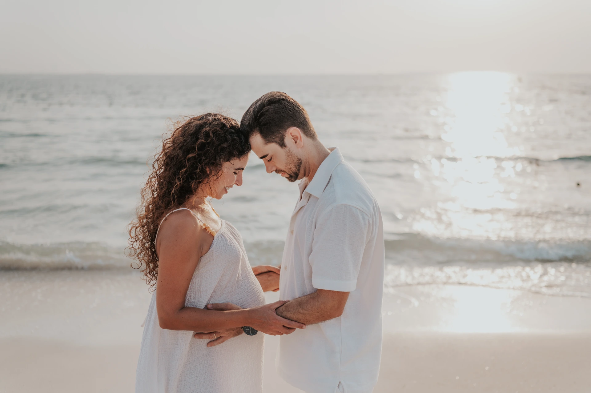 Sunset Dubai Beach Maternity Couple An expectant couple touches foreheads and holds each other beside the sea during a sunlit Dubai beach maternity photoshoot.