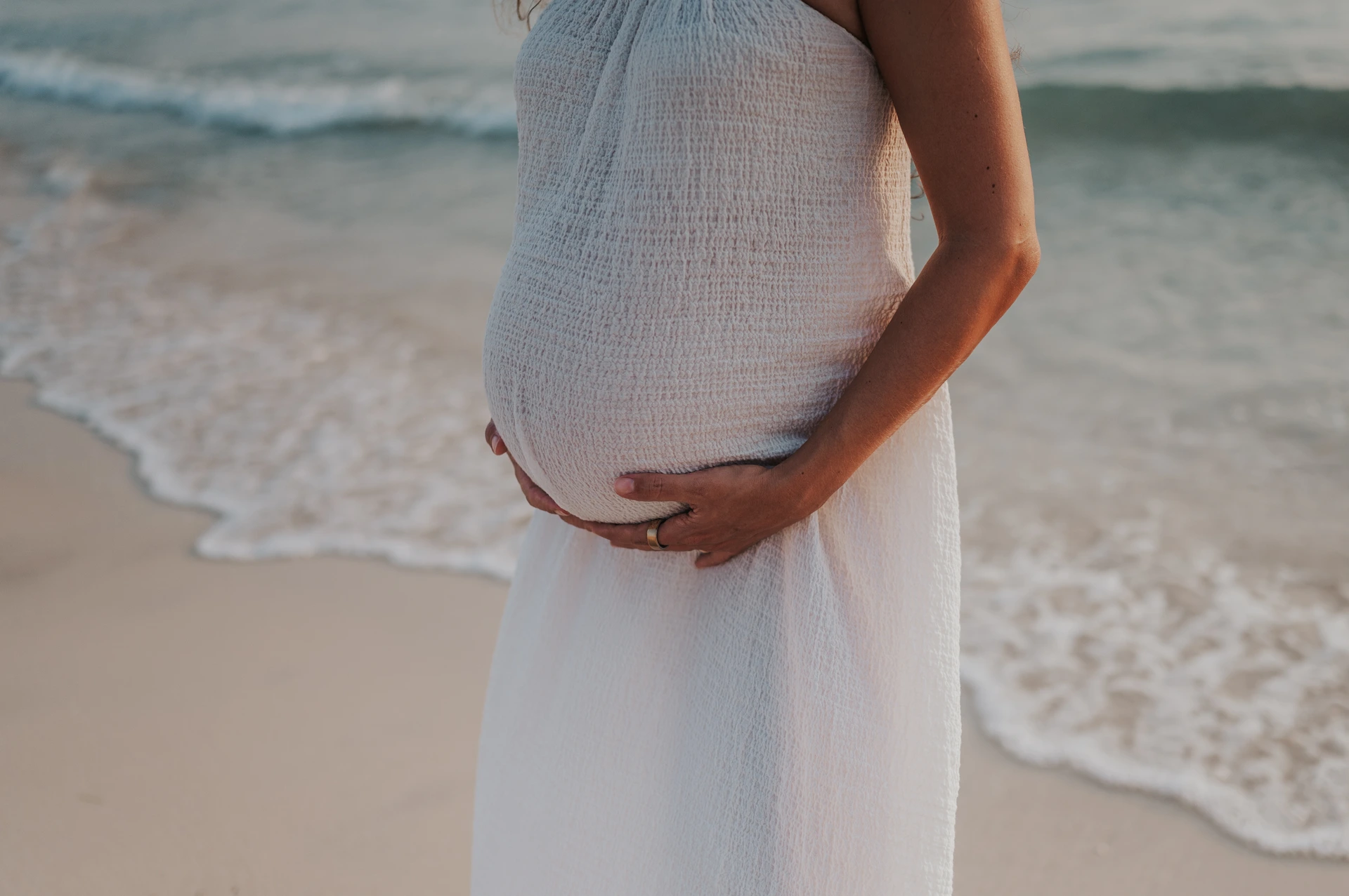 Dubai Beach Maternity Bump by the Waves A pregnant woman in a white dress supports her bump with both hands beside the waves during a Dubai beach maternity session.