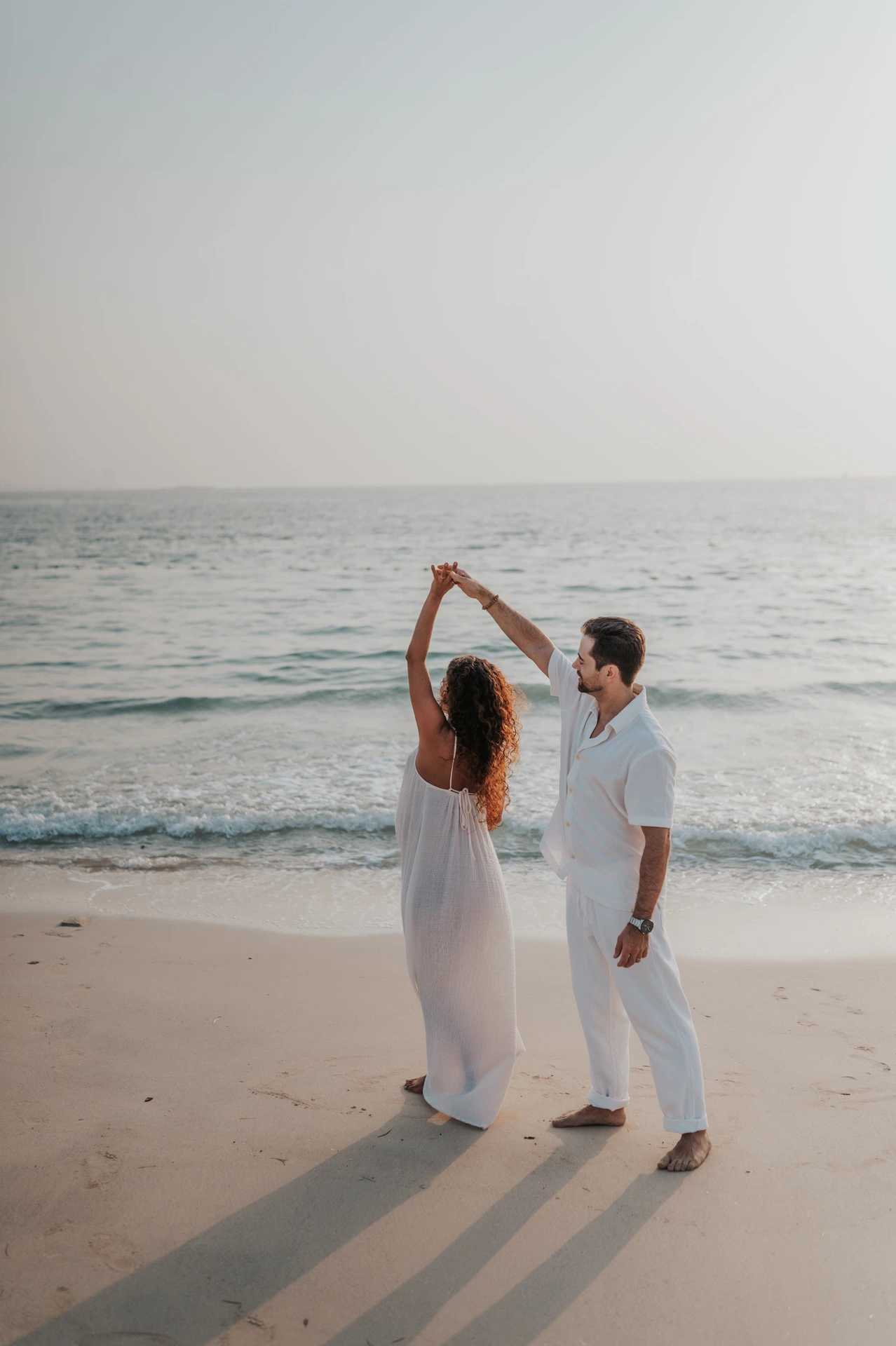 An expectant couple raises joined hands as if dancing beside the sea during a Dubai beach maternity photoshoot.