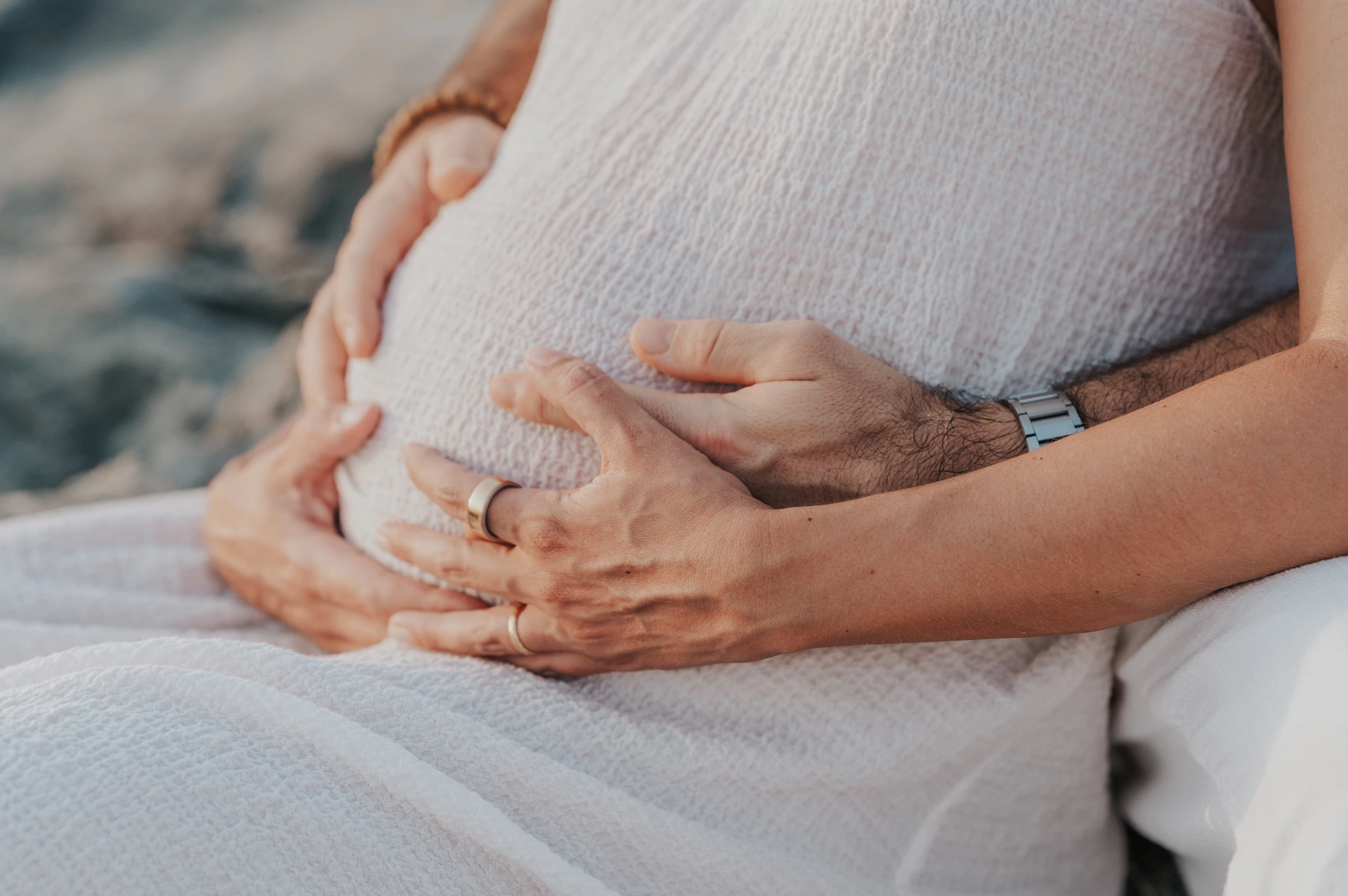 Dubai Beach Maternity Hands Around Bump Two pairs of hands gently cradle a white-covered maternity bump during a close-up Dubai beach portrait.