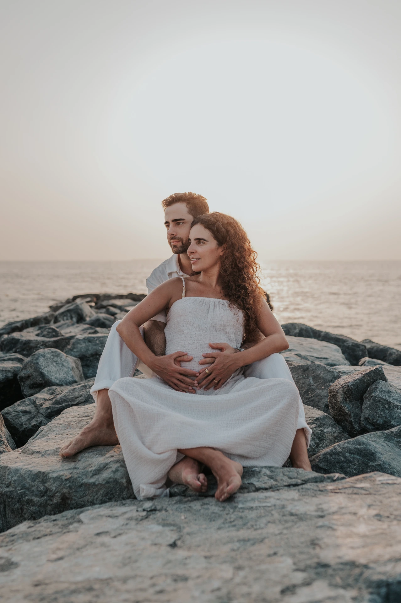 Dubai Coastal Maternity Couple on Rocks An expectant couple sits together on dark shoreline rocks, holding the maternity bump during a Dubai sunset photoshoot.