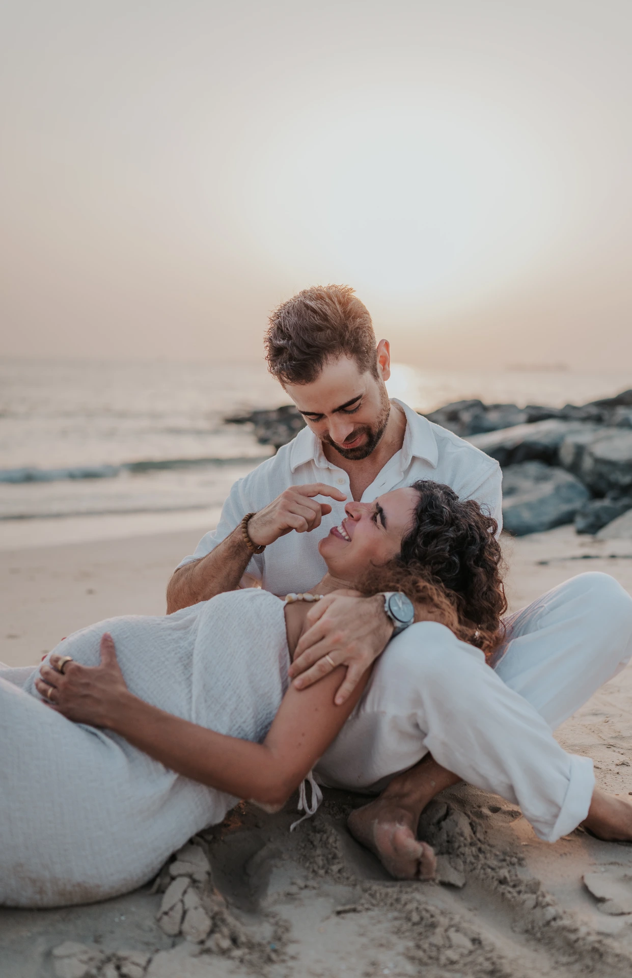 A man playfully touches his pregnant partner's nose as she reclines against him during a Dubai beach maternity session.