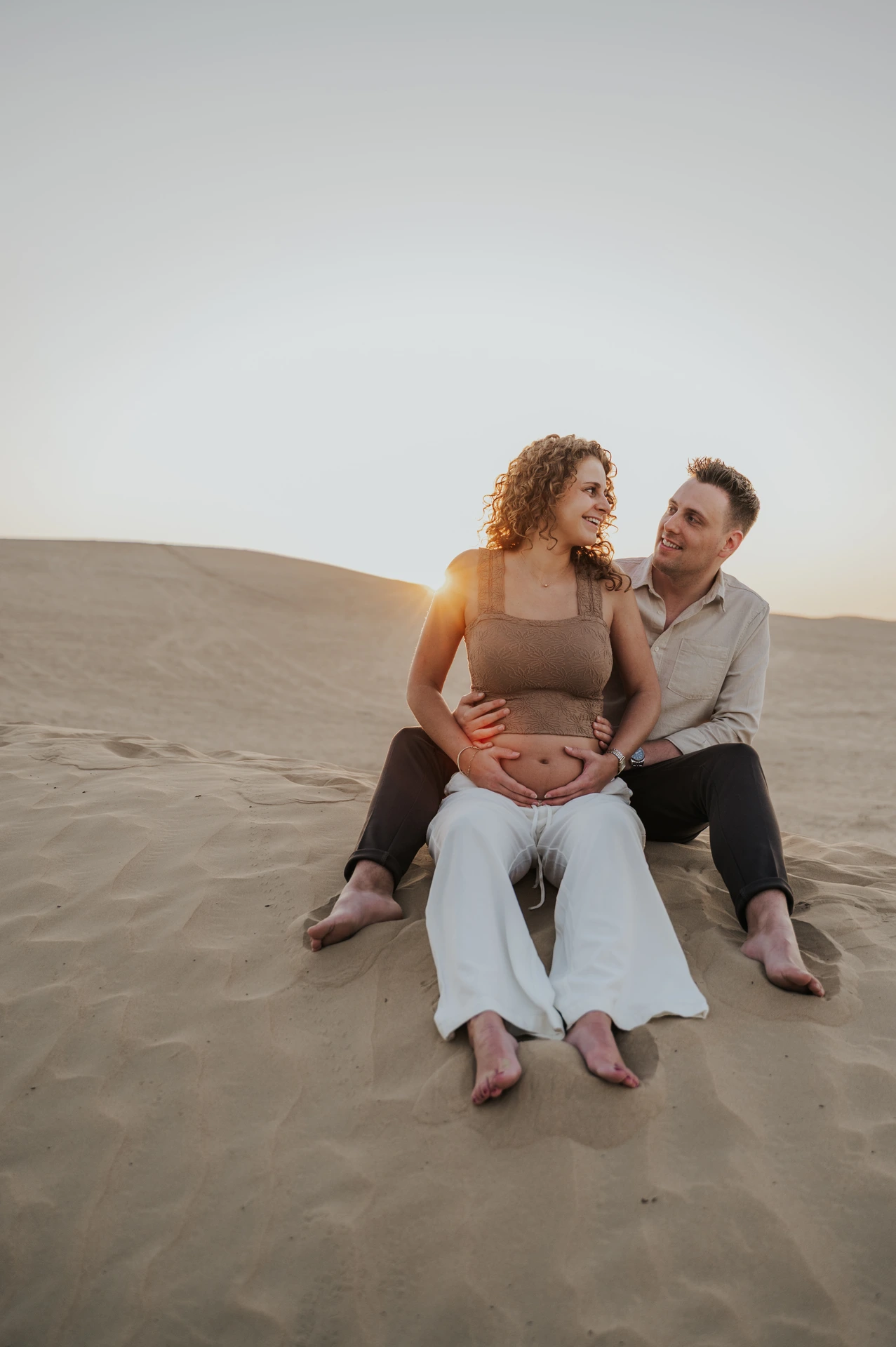An expectant couple sits close together on a Dubai desert dune, smiling at each other during a sunset maternity photoshoot.