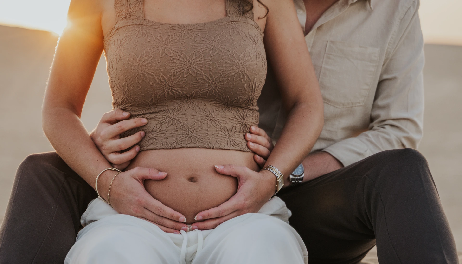 An expectant couple places their hands around a bare bump in warm sunlight during a Dubai desert maternity portrait.