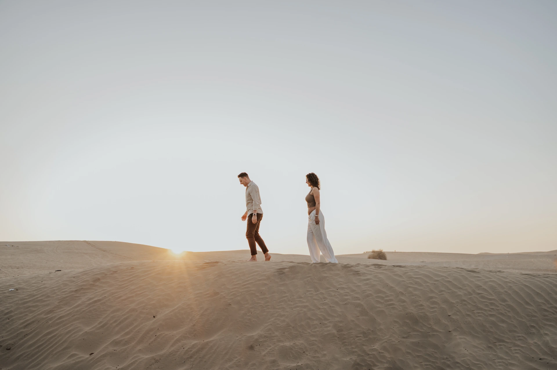 An expectant couple walks barefoot along the crest of a Dubai desert dune during their sunset maternity photoshoot.