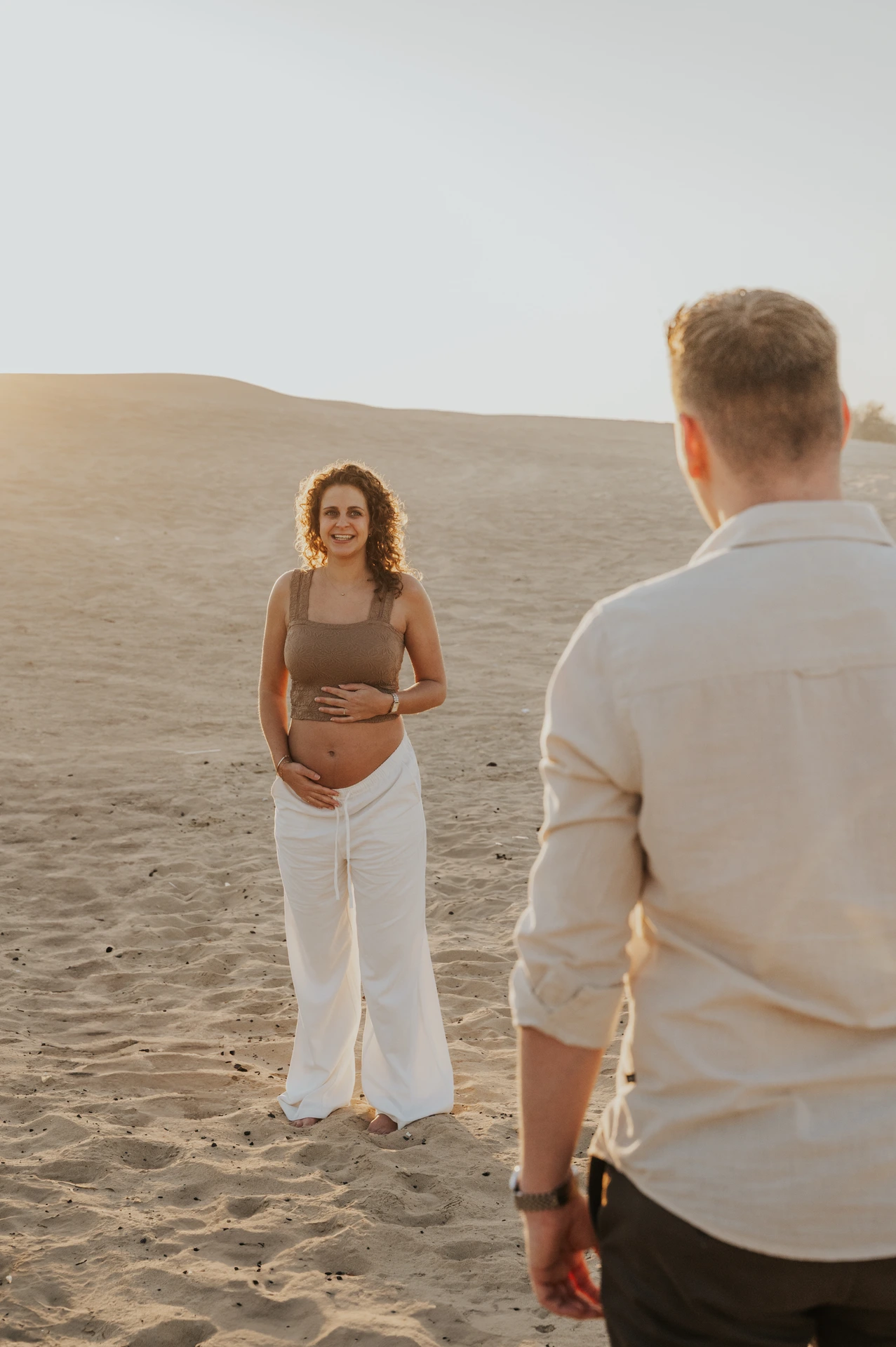 A pregnant woman smiles toward her approaching partner while holding her bump on a sunlit Dubai desert maternity shoot.