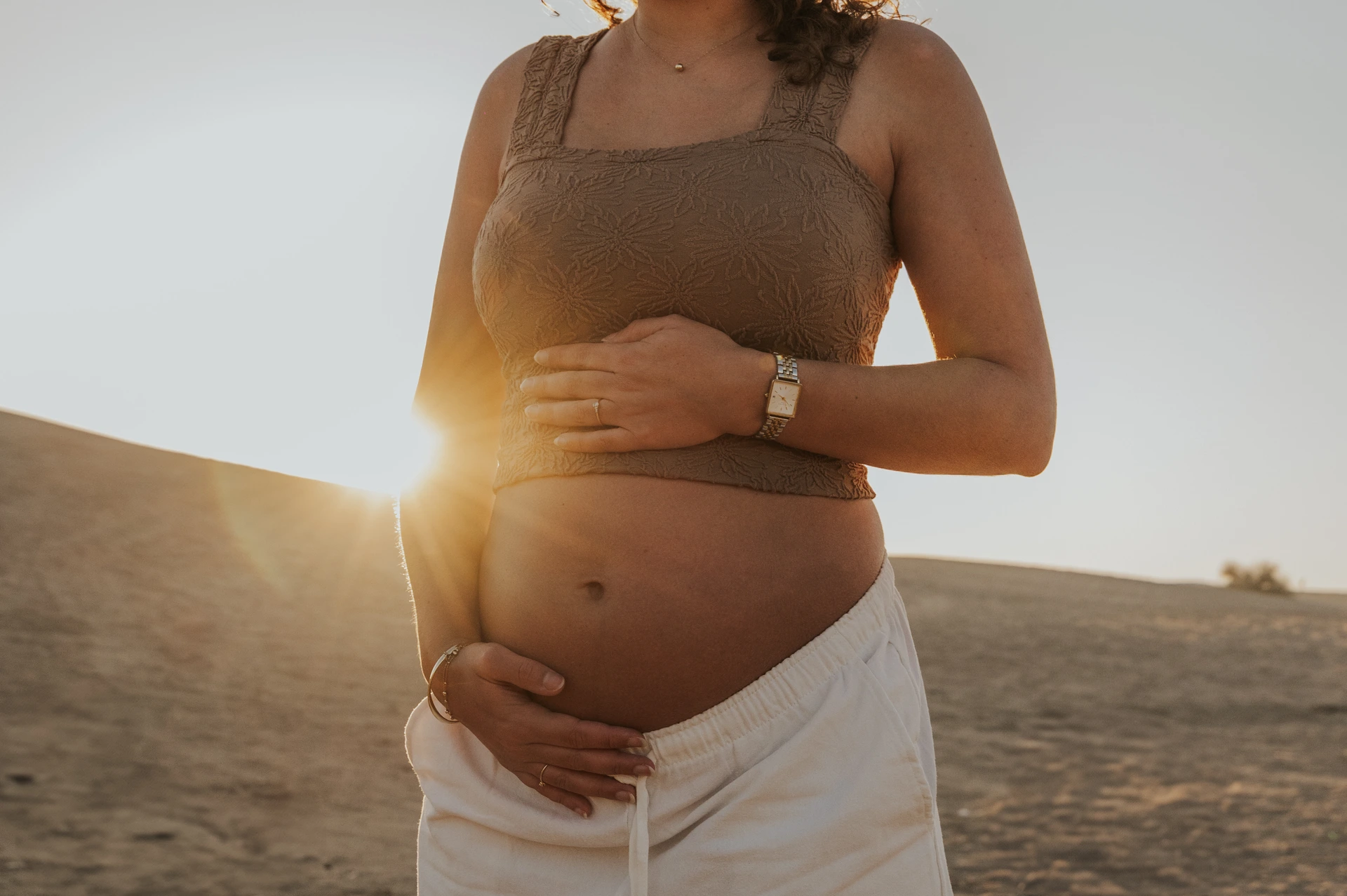 A pregnant woman in a brown top holds her bare bump as sunset crosses the Dubai desert maternity scene.