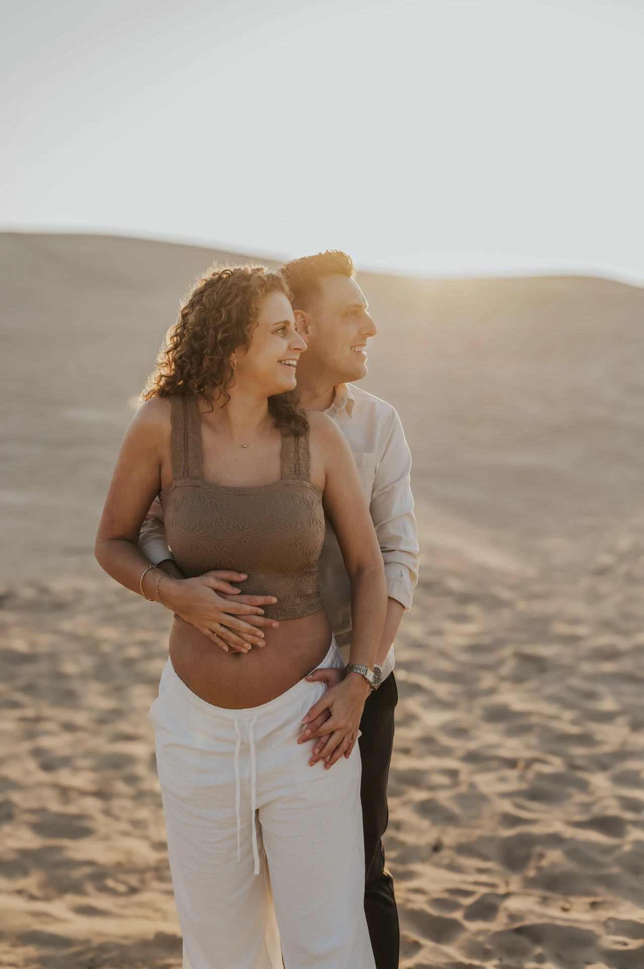 An expectant couple stands together on a Dubai desert dune, smiling into the evening light during their maternity photoshoot.