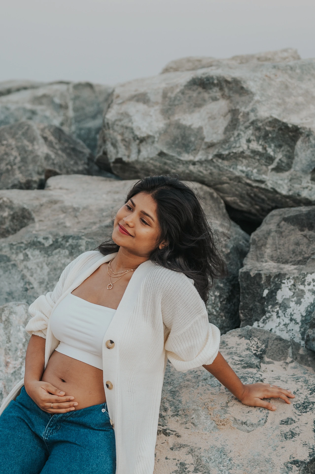 Dubai Coastal Maternity Portrait on Rocks A pregnant woman in denim and a cream cardigan leans against shoreline rocks during a Dubai coastal maternity photoshoot.