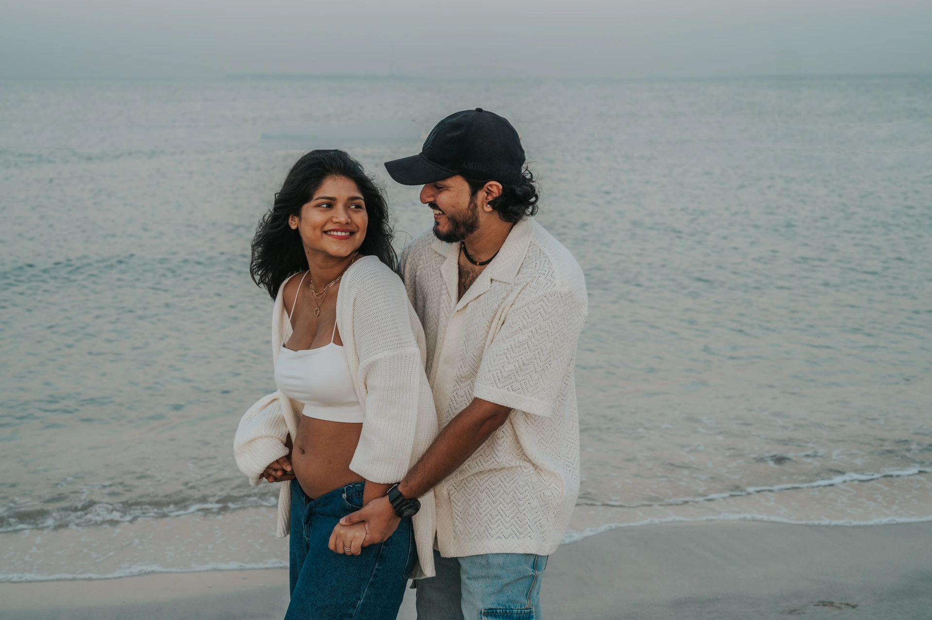 An expectant couple laughs together at the water's edge during a relaxed Dubai beach maternity photoshoot.