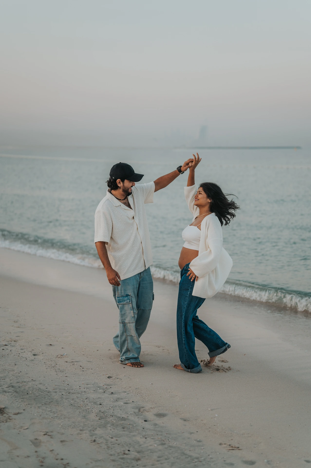 A man lifts his pregnant partner's hand as she turns beneath it during a playful Dubai beach maternity session.