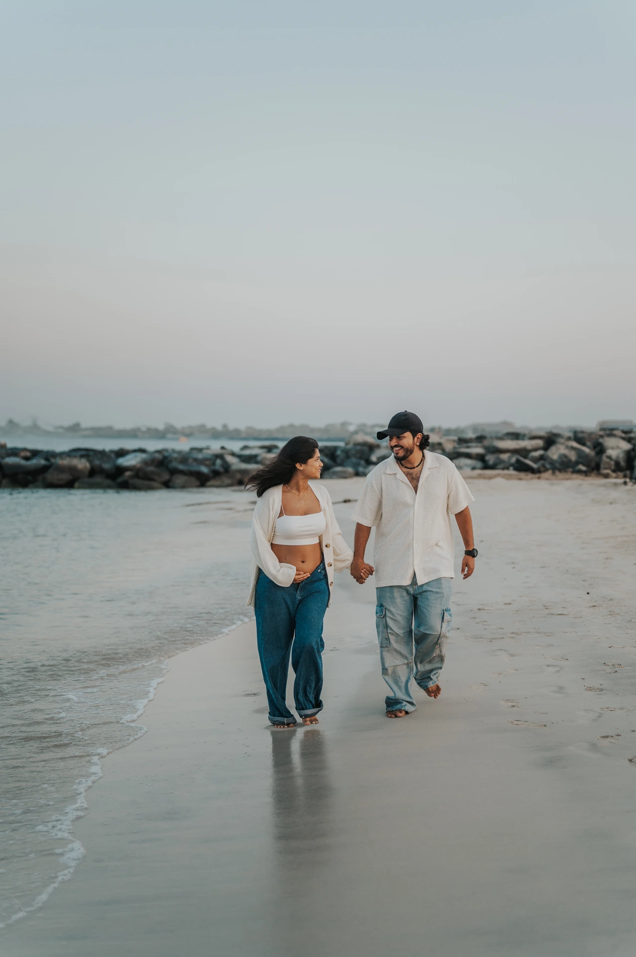 Walking Dubai Beach Maternity Couple An expectant couple walks barefoot hand in hand beside calm water during their Dubai beach maternity photoshoot.