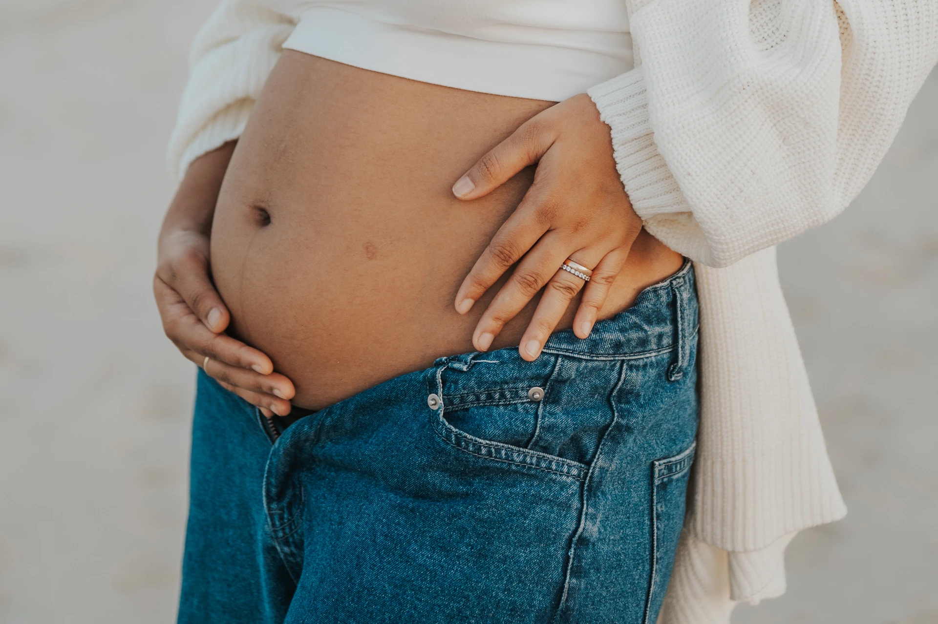 A pregnant woman rests both hands on her bare bump above loose jeans during a close-up Dubai beach maternity portrait.