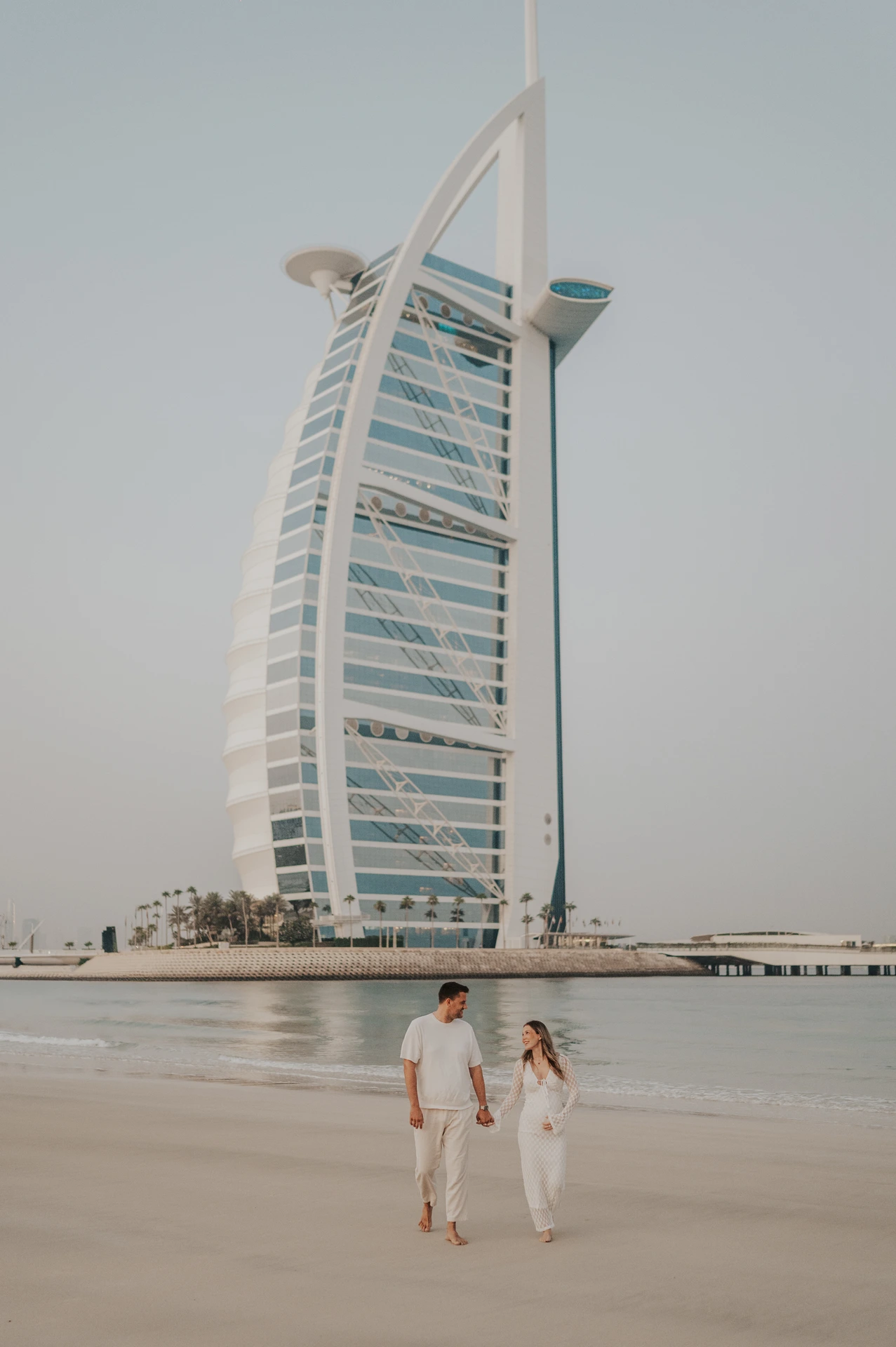 An expectant couple walks barefoot along the shore with Burj Al Arab behind them during a Dubai maternity photoshoot.