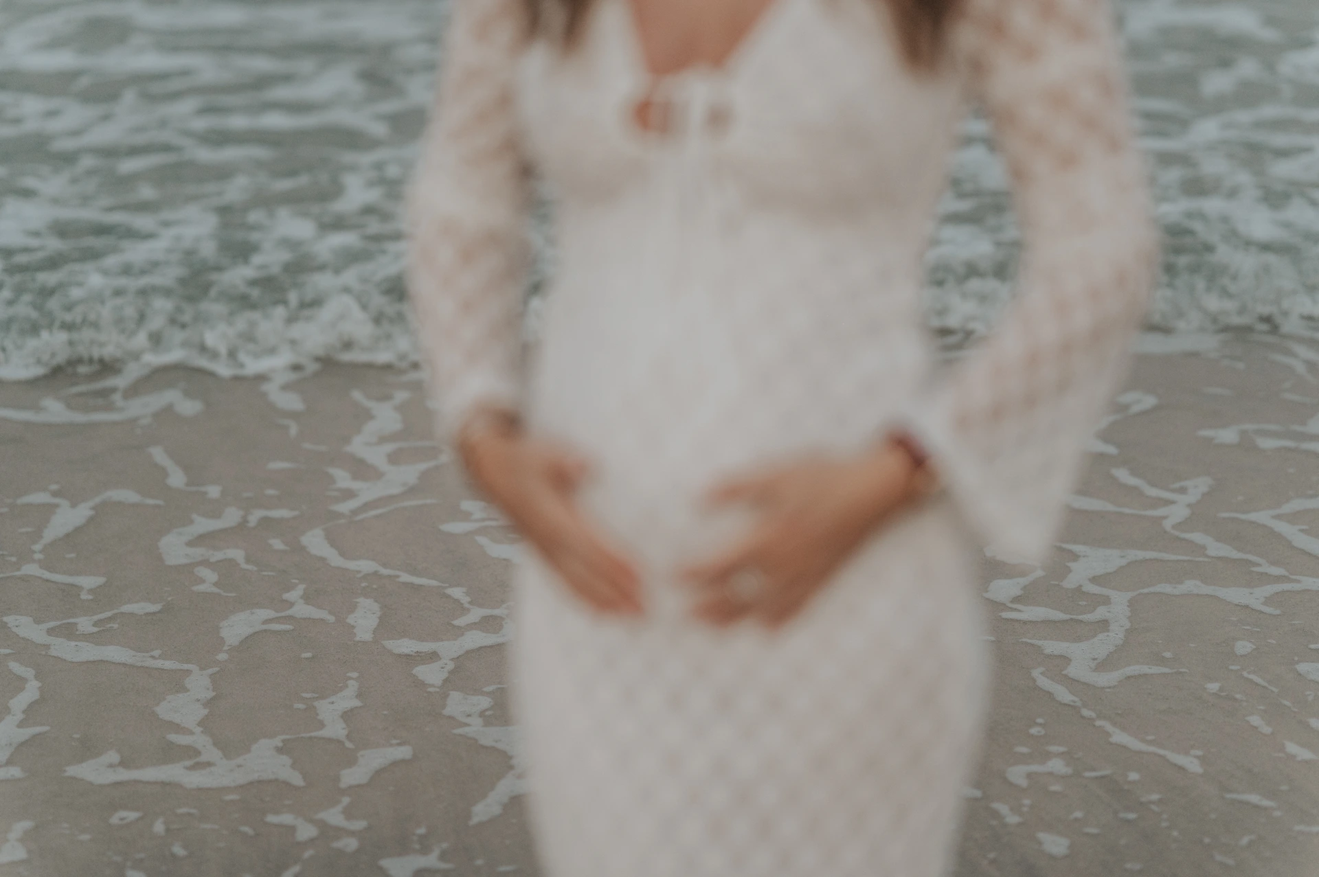 Soft-Focus Dubai Beach Maternity Portrait A pregnant woman holds her lace-covered bump in soft focus as waves reach the shore during a Dubai beach maternity portrait.