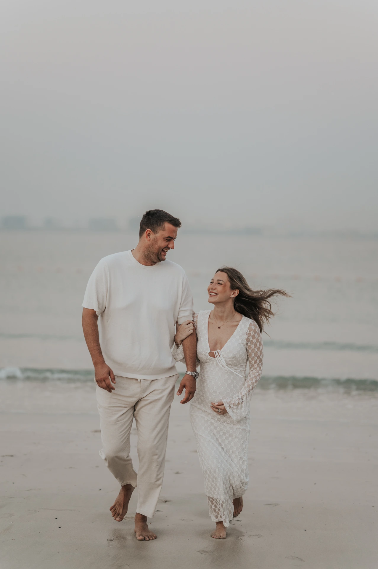 Walking Dubai Beach Maternity Couple in White An expectant couple walks arm in arm along the shoreline in white clothing during a Dubai beach maternity photoshoot.