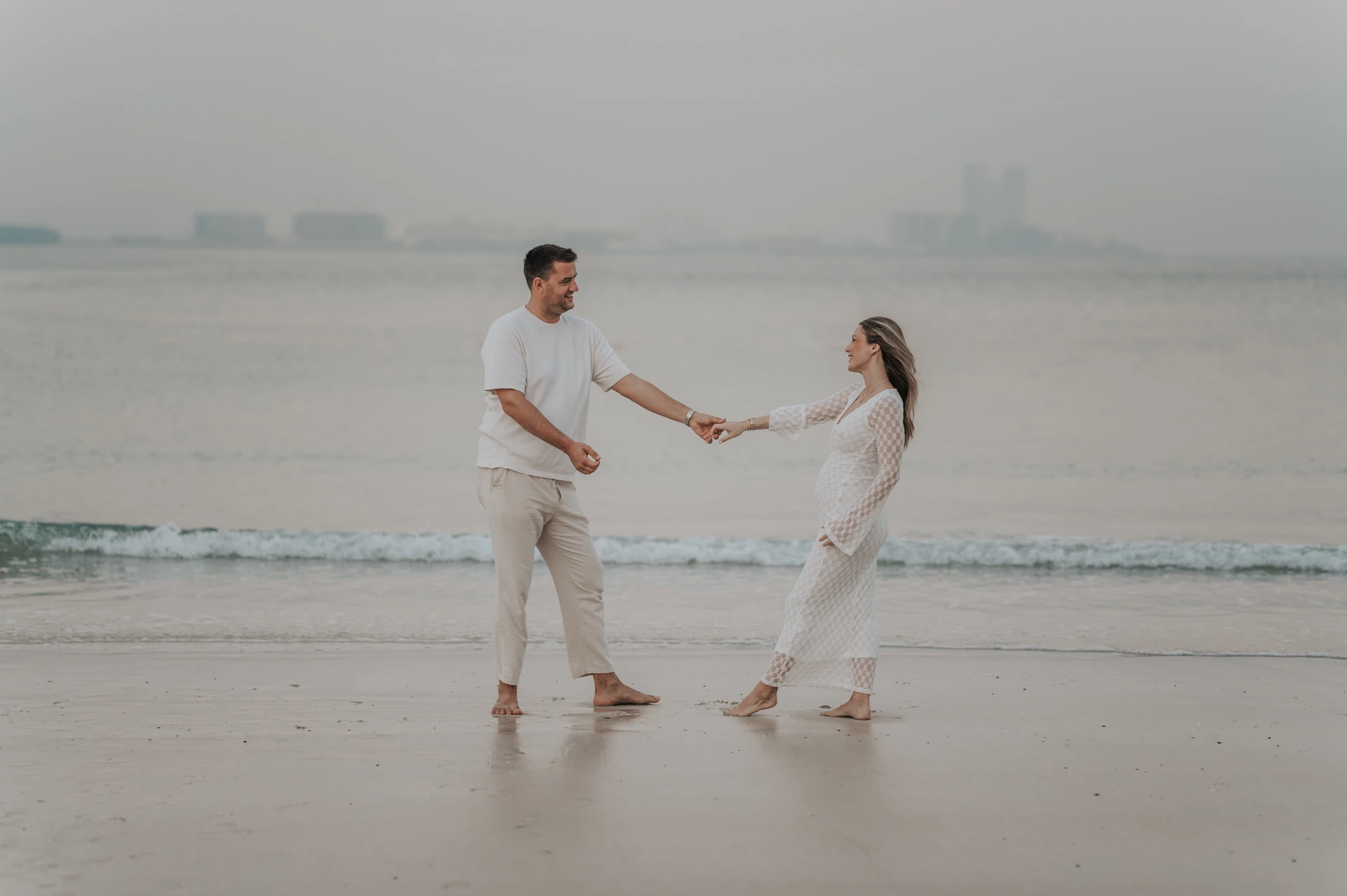 Playful Dubai Beach Maternity Couple Holding Hands An expectant couple smiles while holding hands across the wet sand during a playful Dubai beach maternity session.