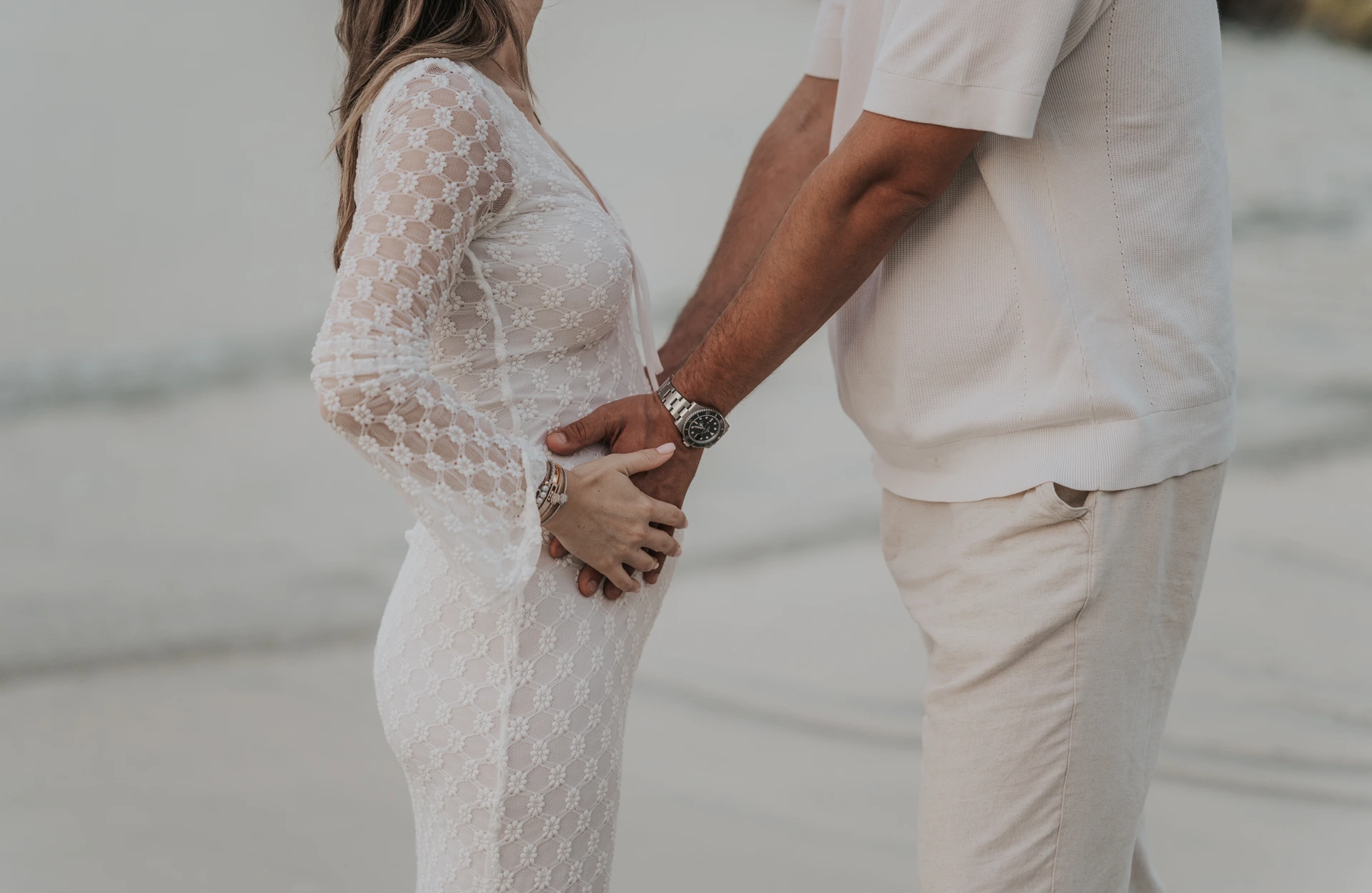 Dubai Beach Maternity Bump With Partner's Hands A man places both hands around his pregnant partner's bump as they face each other during a Dubai beach maternity portrait.