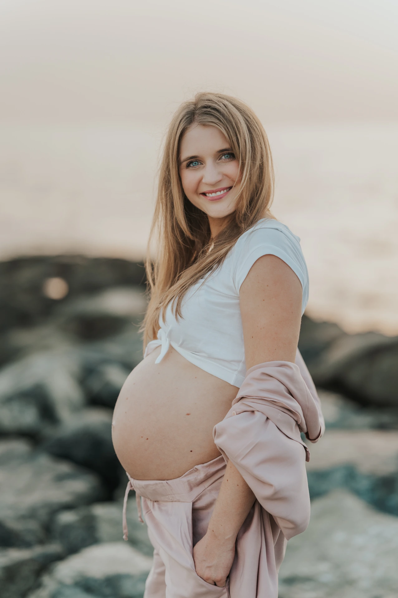 A pregnant woman in blush-pink clothing smiles toward the camera beside shoreline rocks during a Dubai maternity photoshoot.