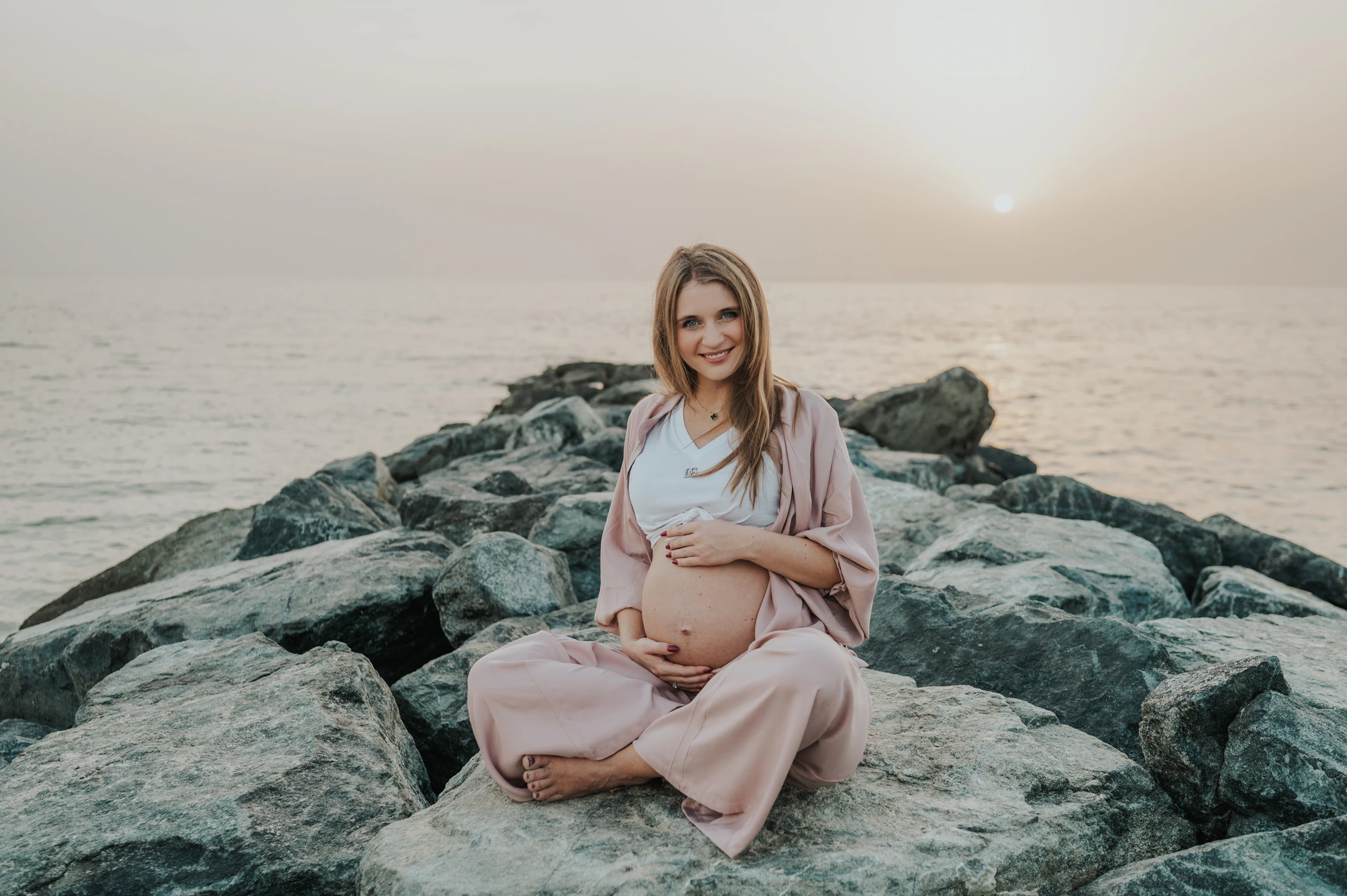 A pregnant woman sits cross-legged on coastal rocks, holding her bump at sunset during a Dubai maternity photoshoot.