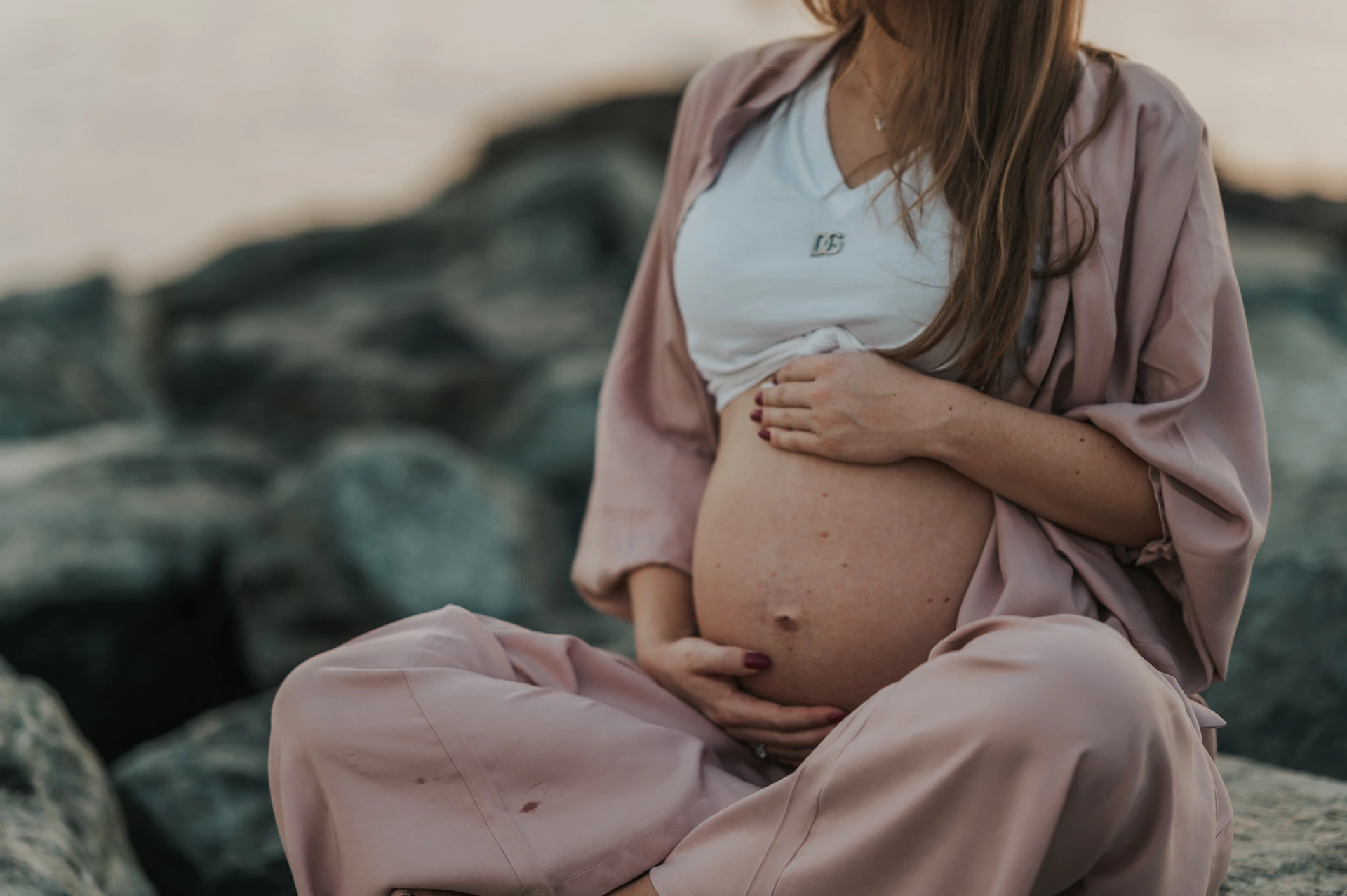 A pregnant woman in blush-pink clothing cradles her bare bump while seated on rocks during a Dubai maternity portrait.