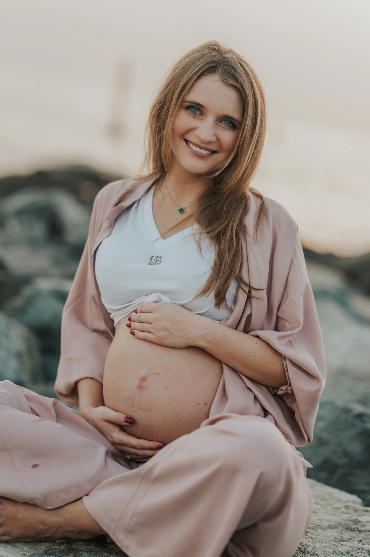 Smiling Dubai Maternity Portrait by the Rocks A pregnant woman in blush-pink clothing smiles and holds her bump while seated on shoreline rocks during a Dubai maternity photoshoot.