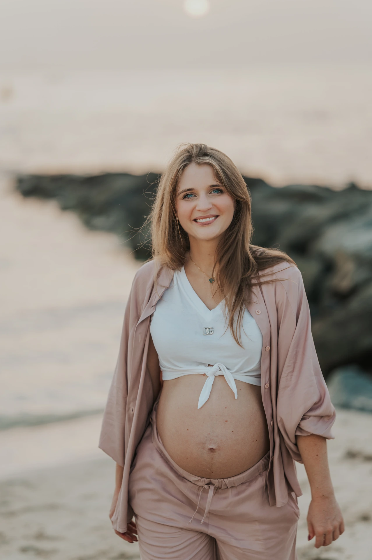 Dubai Beach Maternity Portrait in Evening Light A pregnant woman in blush-pink clothing stands smiling near shoreline rocks during a softly lit Dubai beach maternity portrait.