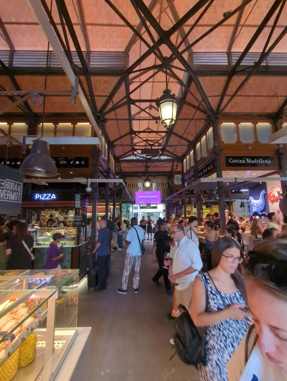Visitors browse food stalls beneath iron roof trusses and warm lanterns inside Mercado de San Miguel during this Madrid route.