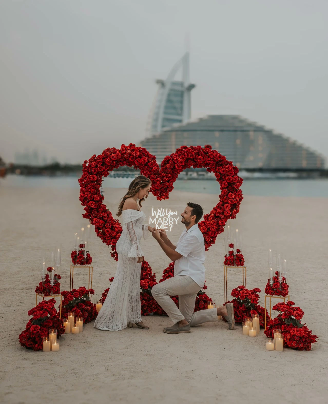 A man kneels to propose on Dubai beach with a red floral heart setup, with Burj Al Arab in the background during a proposal photoshoot.