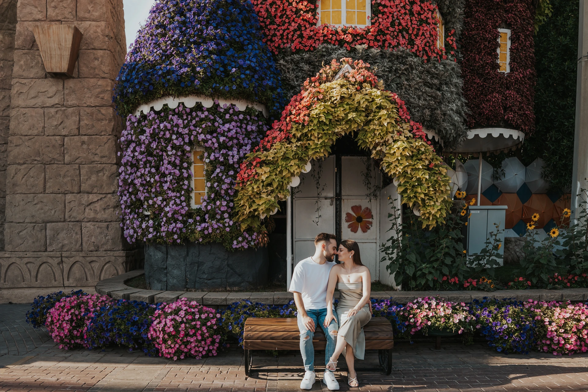 A couple sits together on a bench beside a flower-covered cottage in Dubai Miracle Garden during a couple photoshoot.