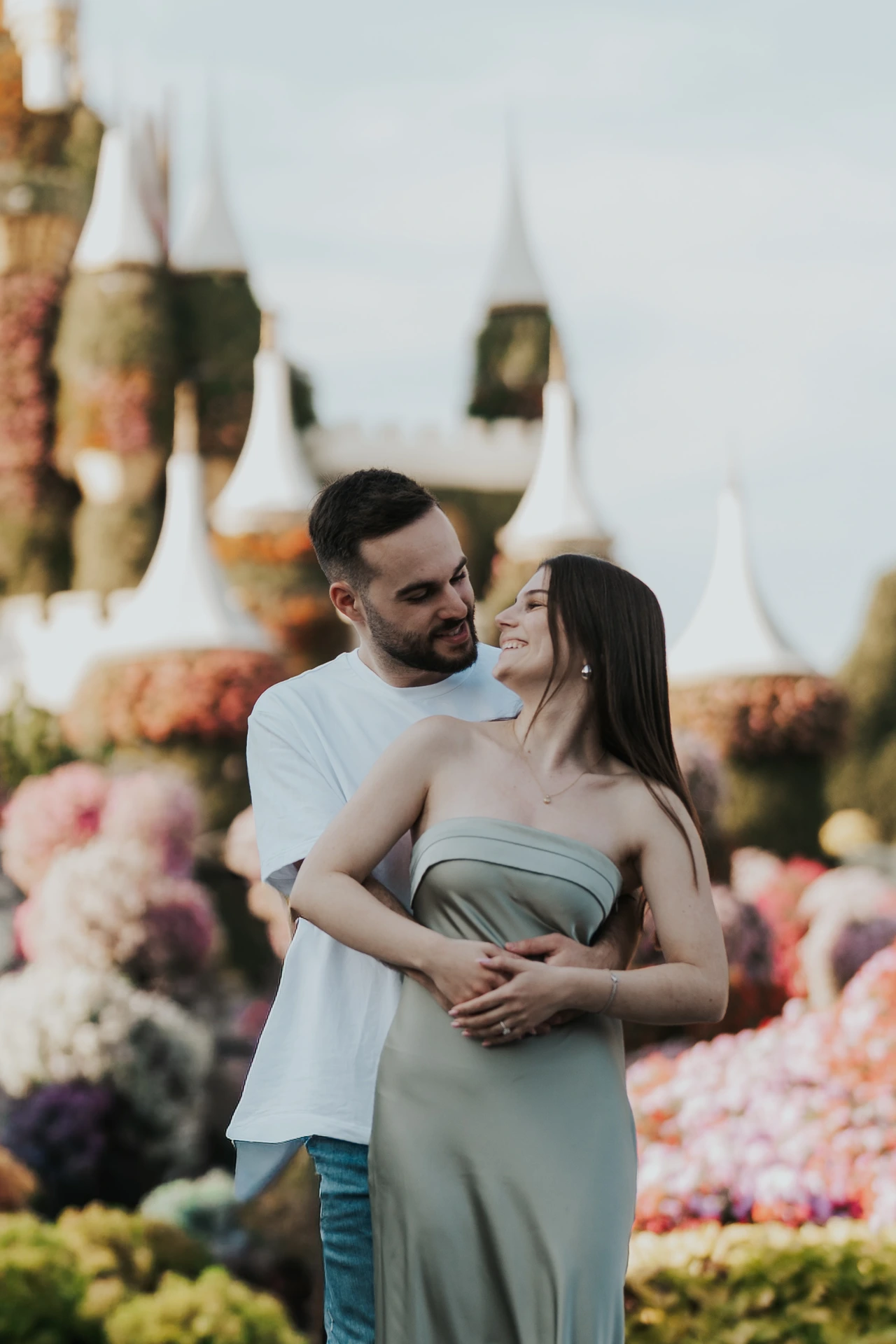 A couple is smiling at each other with soft floral bokeh in the background at Dubai Miracle Garden during a romantic photoshoot.