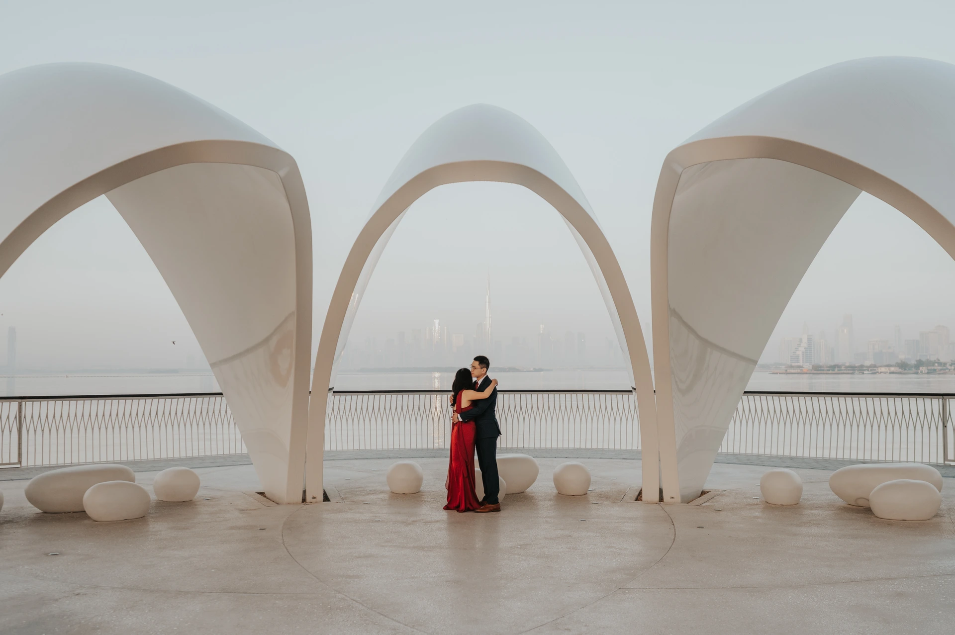 A couple stands under a modern architectural landmark in Dubai with water and skyline views in the background.
