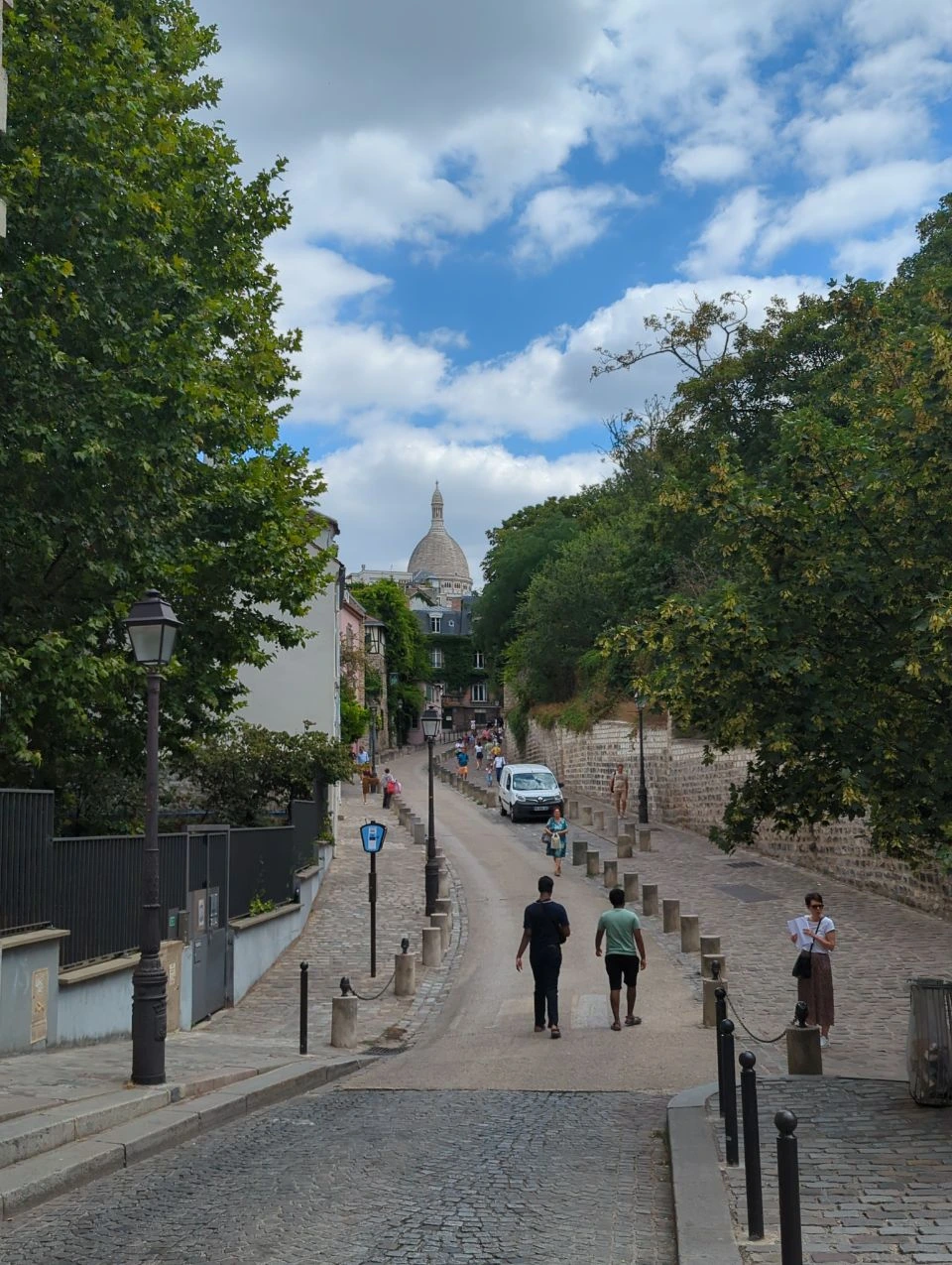 Paris Photo Spots - Montmartre Walking Route A cobbled Montmartre street leads towards the Sacre-Coeur dome in Paris, with pedestrians showing a relaxed photoshoot route on foot.
