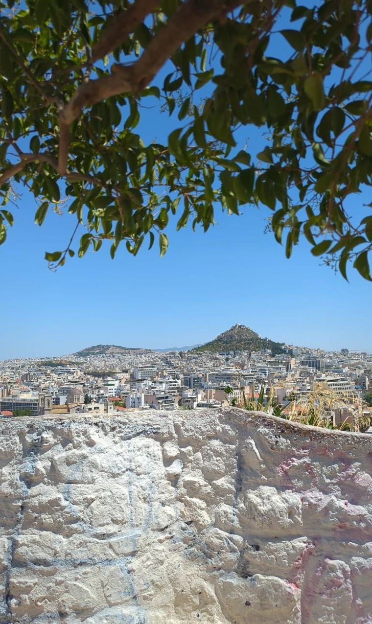 Acropolis Viewpoints - Mount Lycabettus Mount Lycabettus rises above the Athens skyline, framed by leafy branches and a pale stone wall.