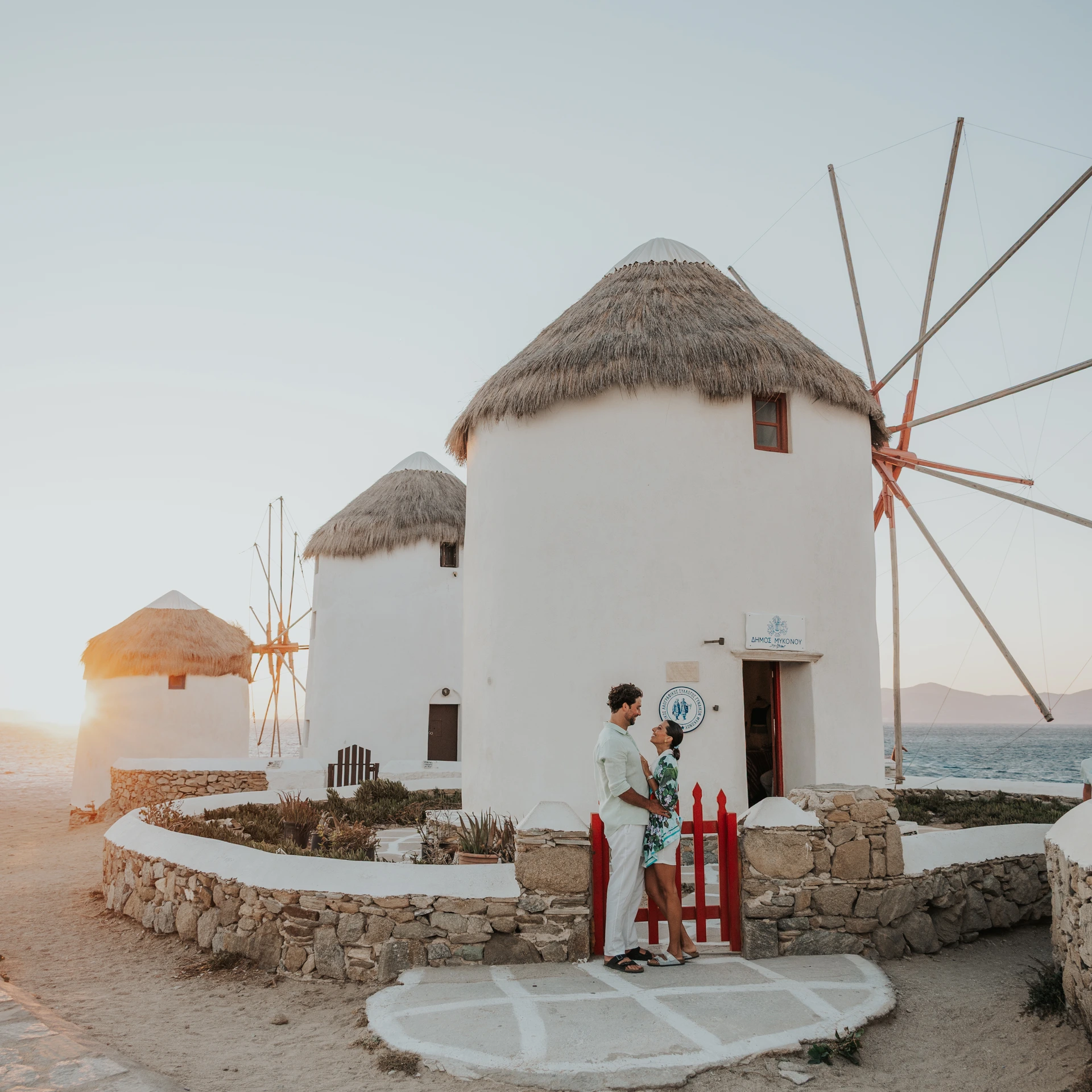 Two adults embrace beside white windmills in Mykonos, with warm low sunlight across the scene.