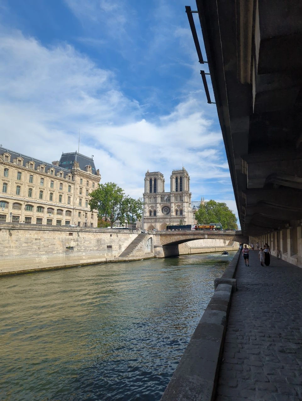 Paris Photo Spots - Notre-Dame Riverside Walk Notre-Dame appears beyond the Seine from a shaded riverside walkway in Paris, offering a layered photoshoot backdrop beside the water.