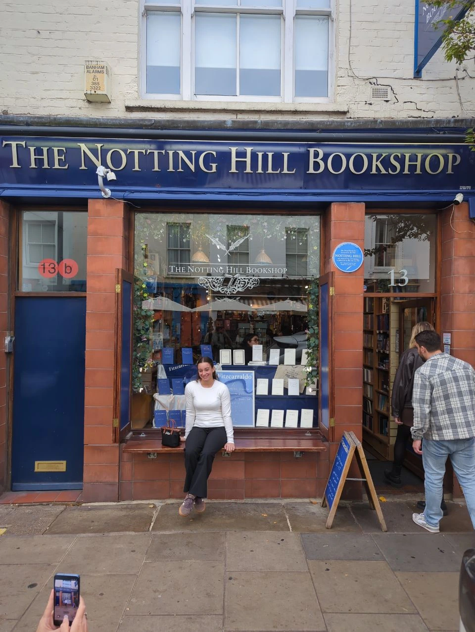 Blue frontage of The Notting Hill Bookshop at number 13, with a visitor seated on the bench and shoppers at the door