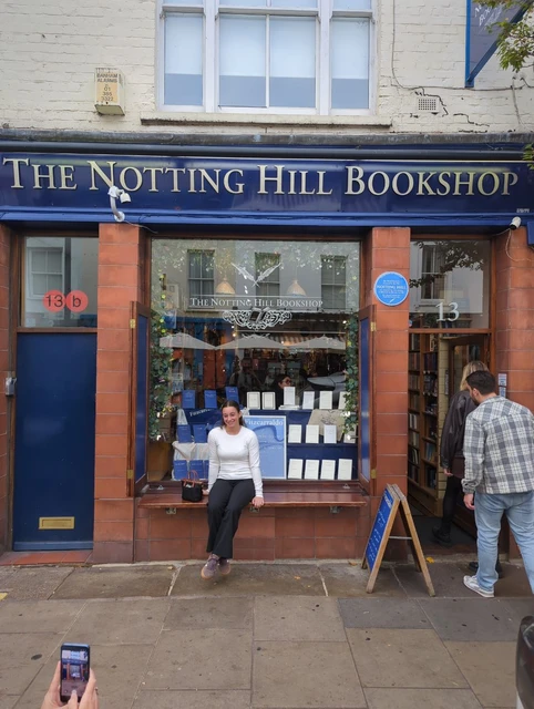 Blue frontage of The Notting Hill Bookshop at number 13, with a visitor seated on the bench and shoppers at the door