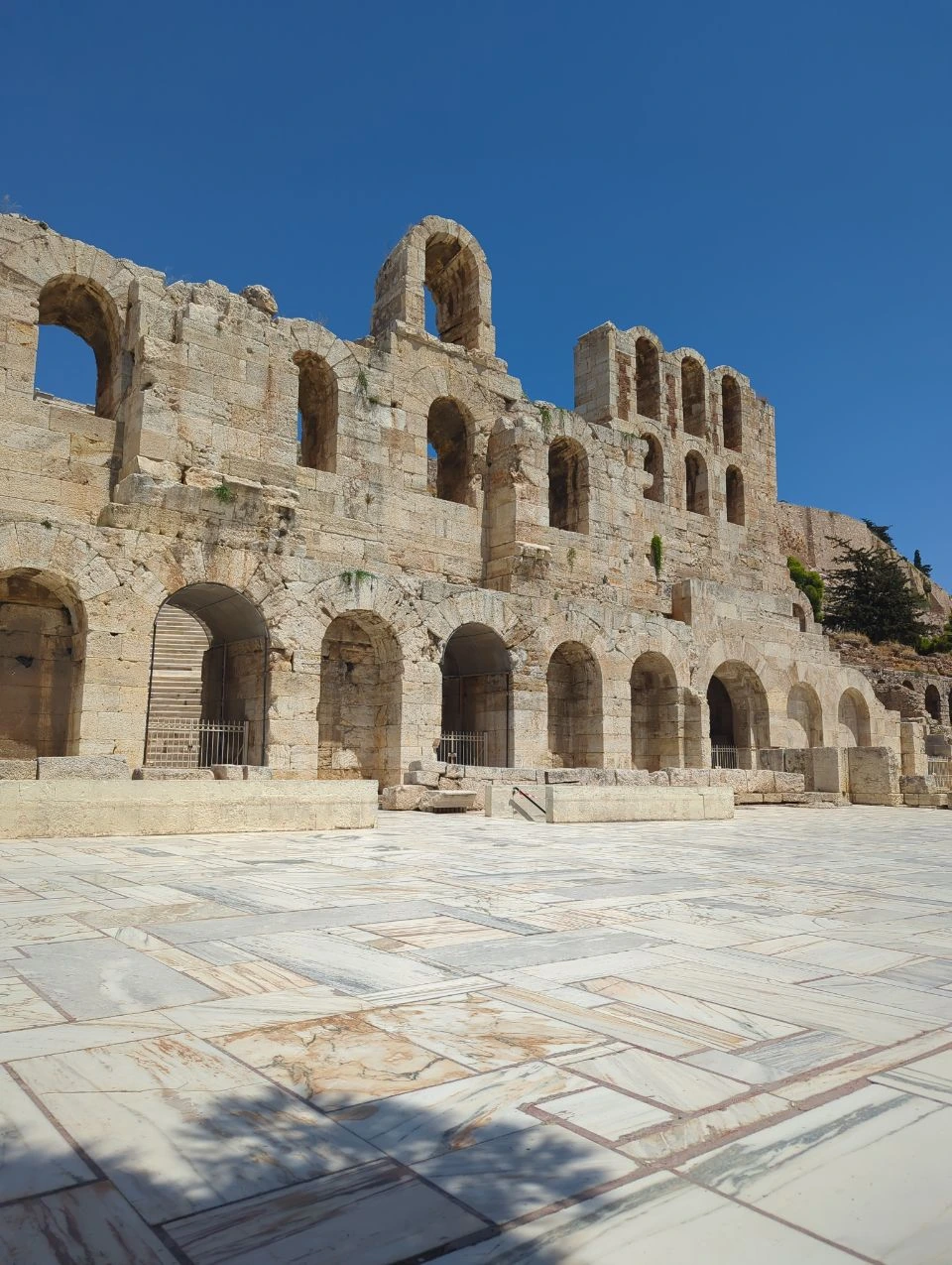 The stone arches of the Odeon of Herodes Atticus rise beside an empty marble courtyard under a clear blue sky.