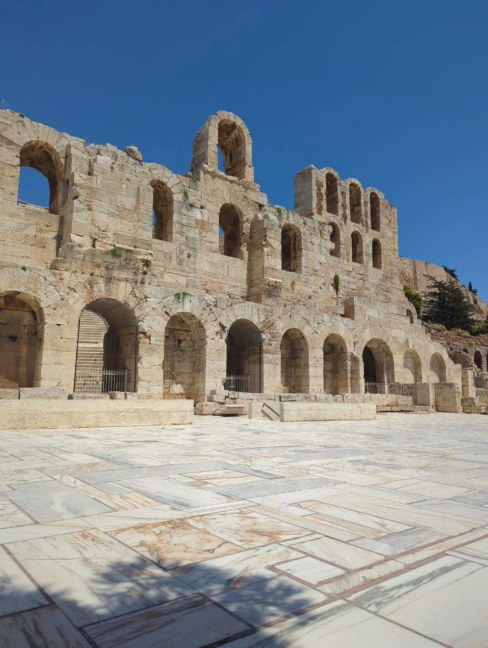 Acropolis Photography Rules - Odeon Architecture The stone arches of the Odeon of Herodes Atticus rise beside an empty marble courtyard under a clear blue sky.