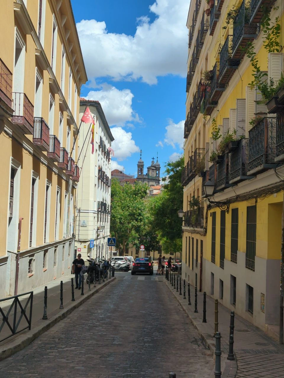 A cobbled street descends between yellow and cream facades in Old Madrid, showing a proposal walk with balconies and distant church towers.