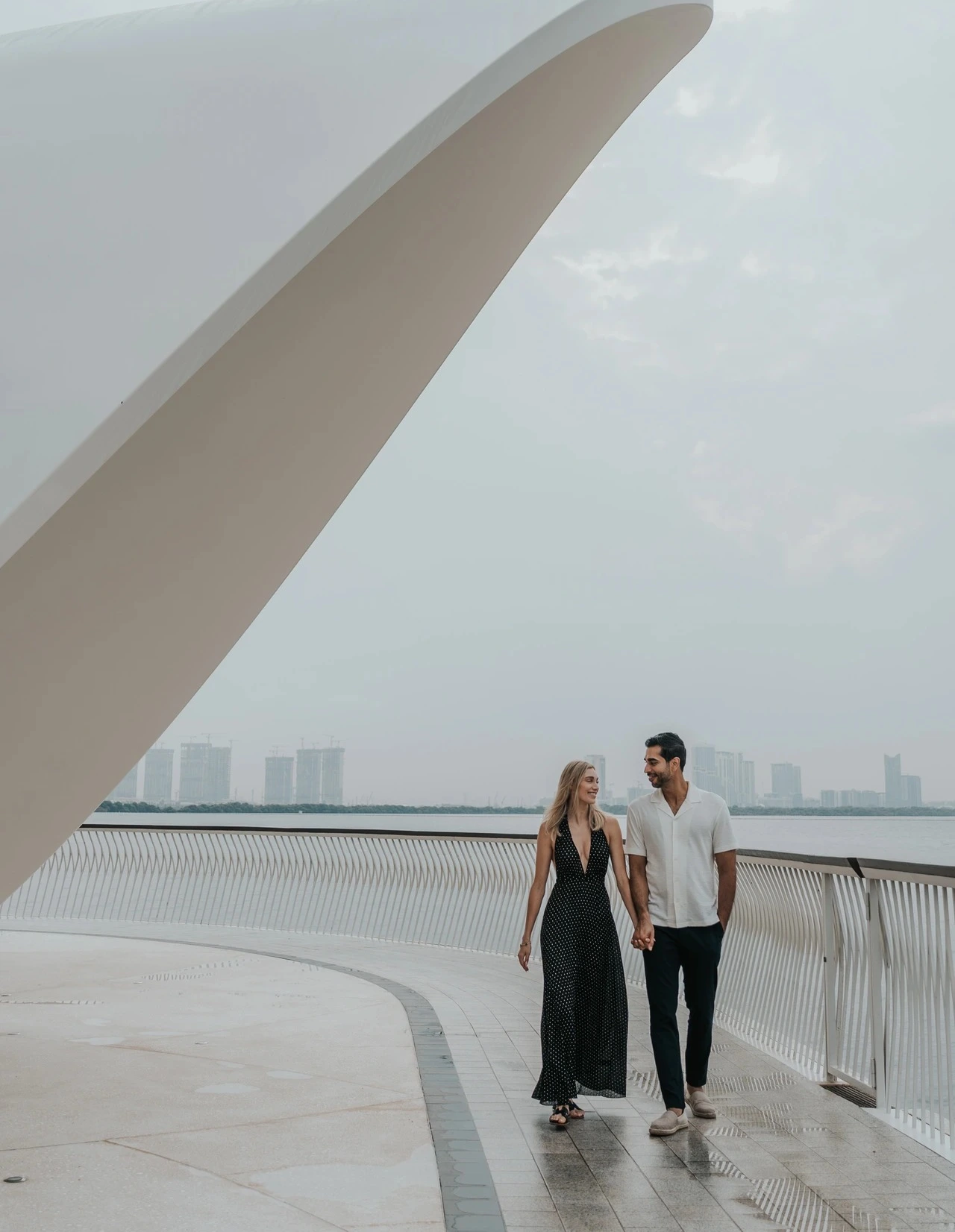 A couple walks along a waterfront promenade with modern architectural frames and skyline views in Dubai.