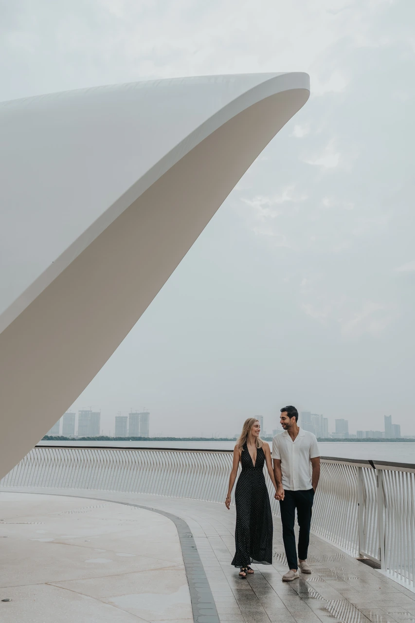 Indoor vs outdoor couple photoshoot dubai - Skyline promenade walk A couple walks side by side along a Dubai waterfront with skyline buildings in the background.