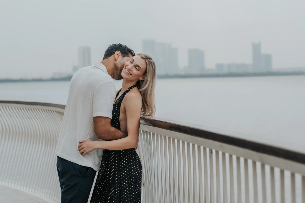 Indoor vs outdoor couple photoshoot dubai - Waterfront railing embrace A couple hugs by a waterfront railing with a soft Dubai skyline backdrop during an outdoor photoshoot.