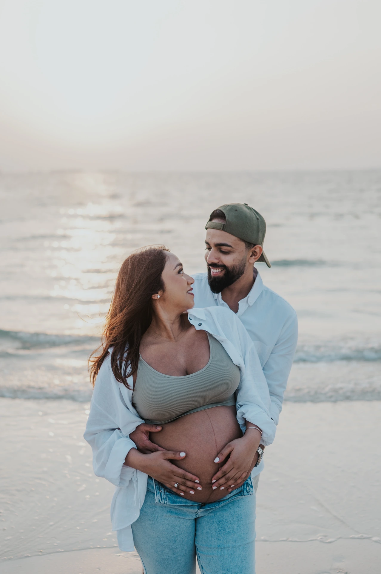 A couple embraces at the Dubai shoreline during a maternity shoot, styled in casual denim and open white layers for a relaxed beach outfit feel.